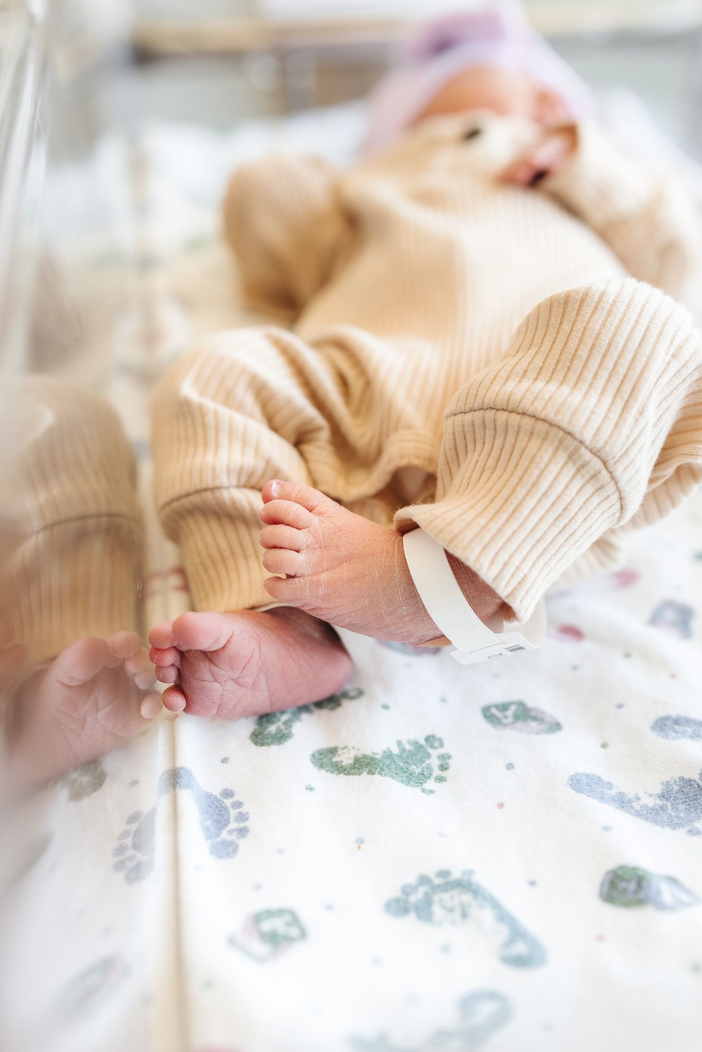 Newborn baby in beige ribbed onesie lying in hospital bassinet, tiny feet visible on footprint patterned blanket.