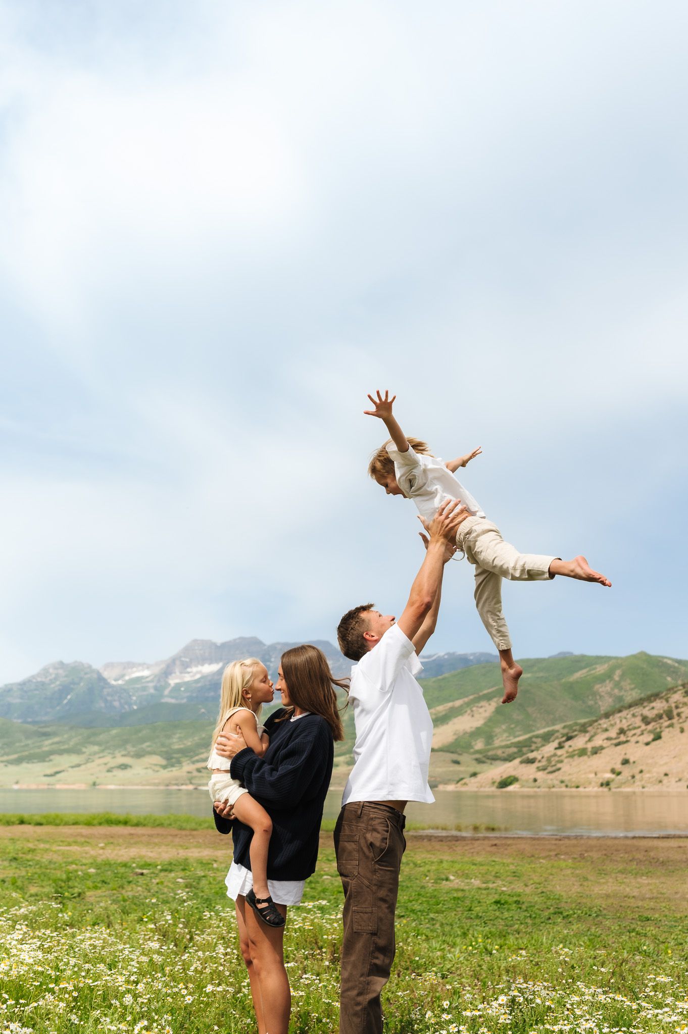 A joyful family outdoors, with a man tossing a child in the air while a woman holds another child in a scenic meadow.