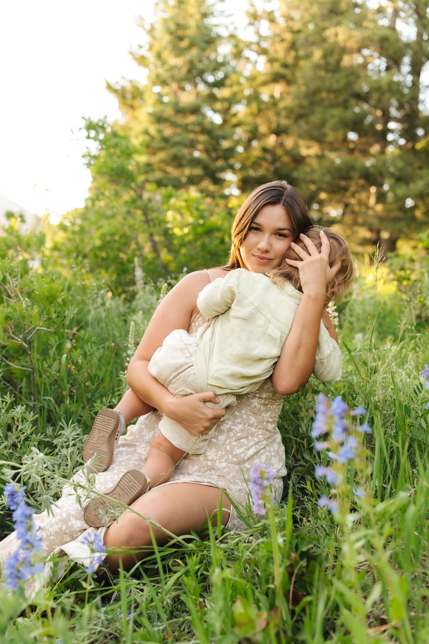 A mother sits in a green meadow with wildflowers, tenderly holding her young child in a warm outdoor setting.
