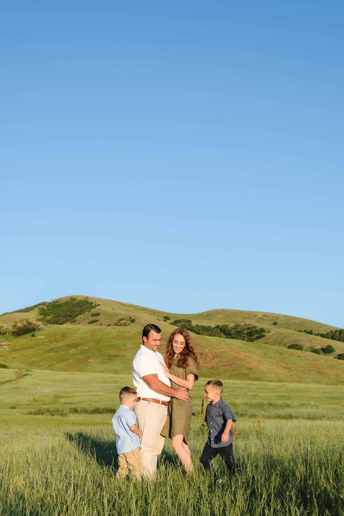 Family of four enjoying a sunny day in a green meadow with rolling hills, parents embracing while two boys play nearby.