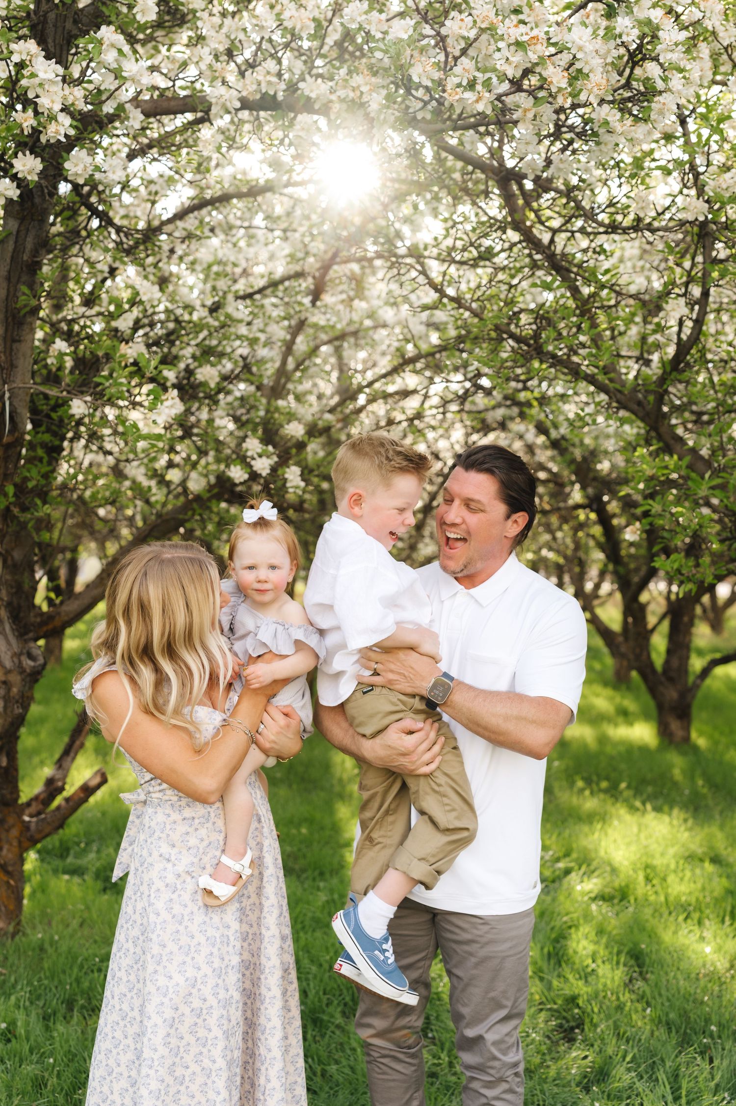 Family of four smiling among blooming white flowering trees in a sunny green orchard during spring.