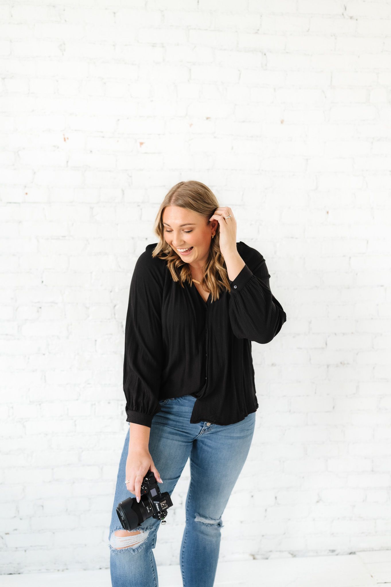Fashion photographer in black shirt and jeans poses playfully against white brick wall with camera.