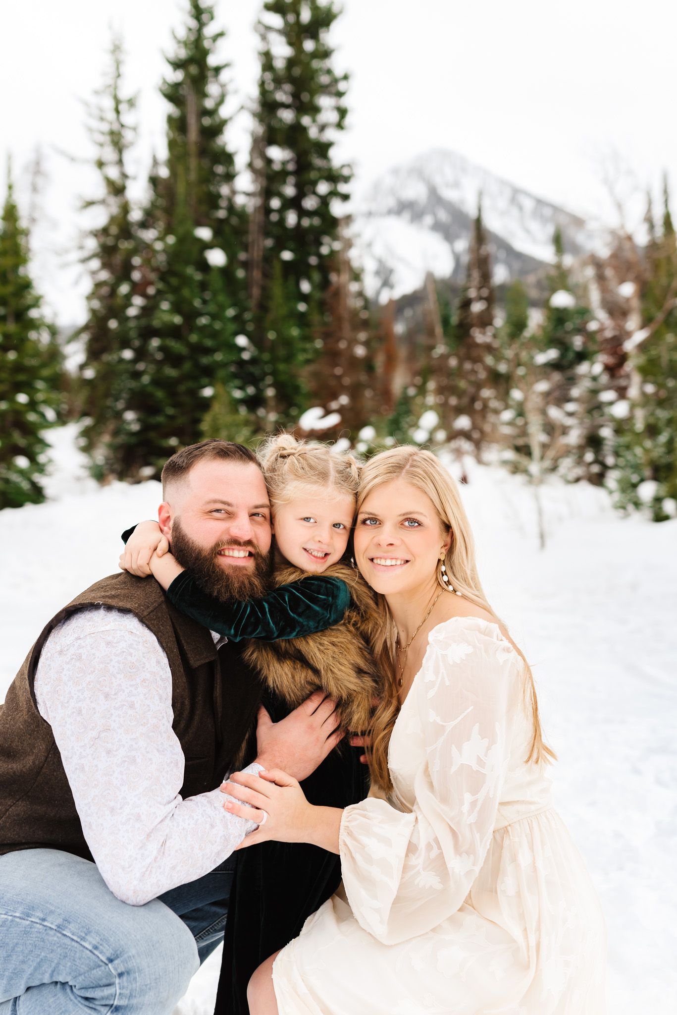 Family sharing an intimate moment together while sitting in deep snow surrounded by pine trees.