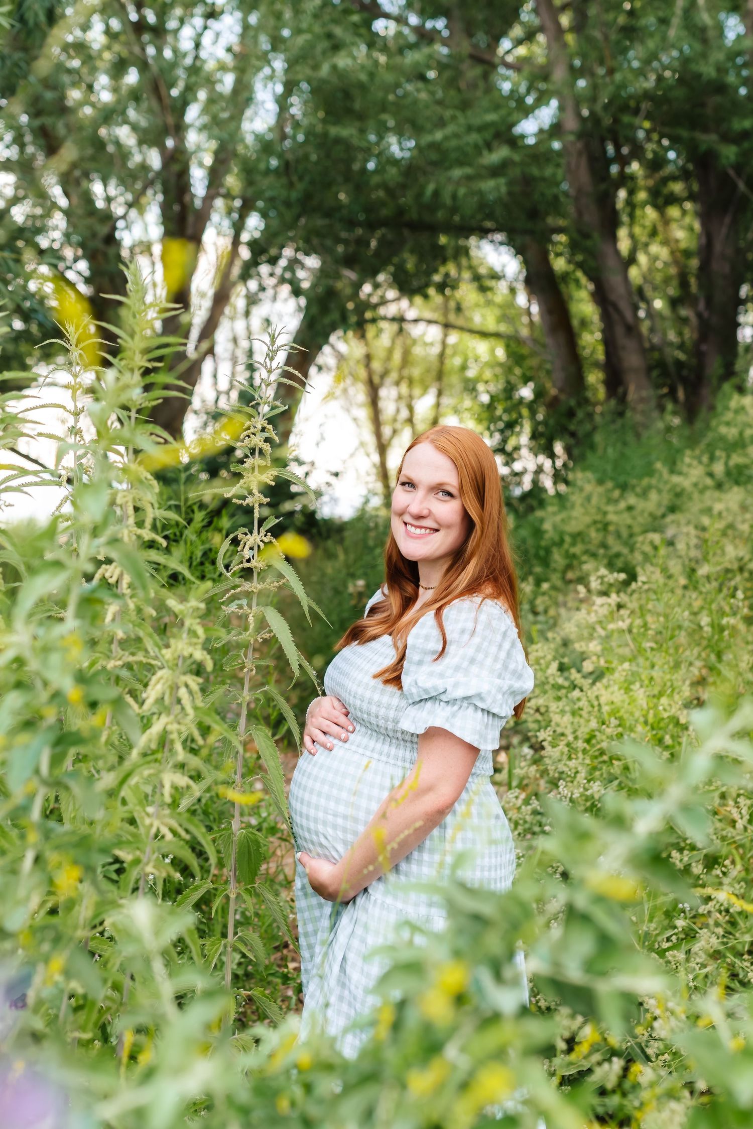 Maternity photo of a woman in a gingham dress standing among wildflowers and greenery.