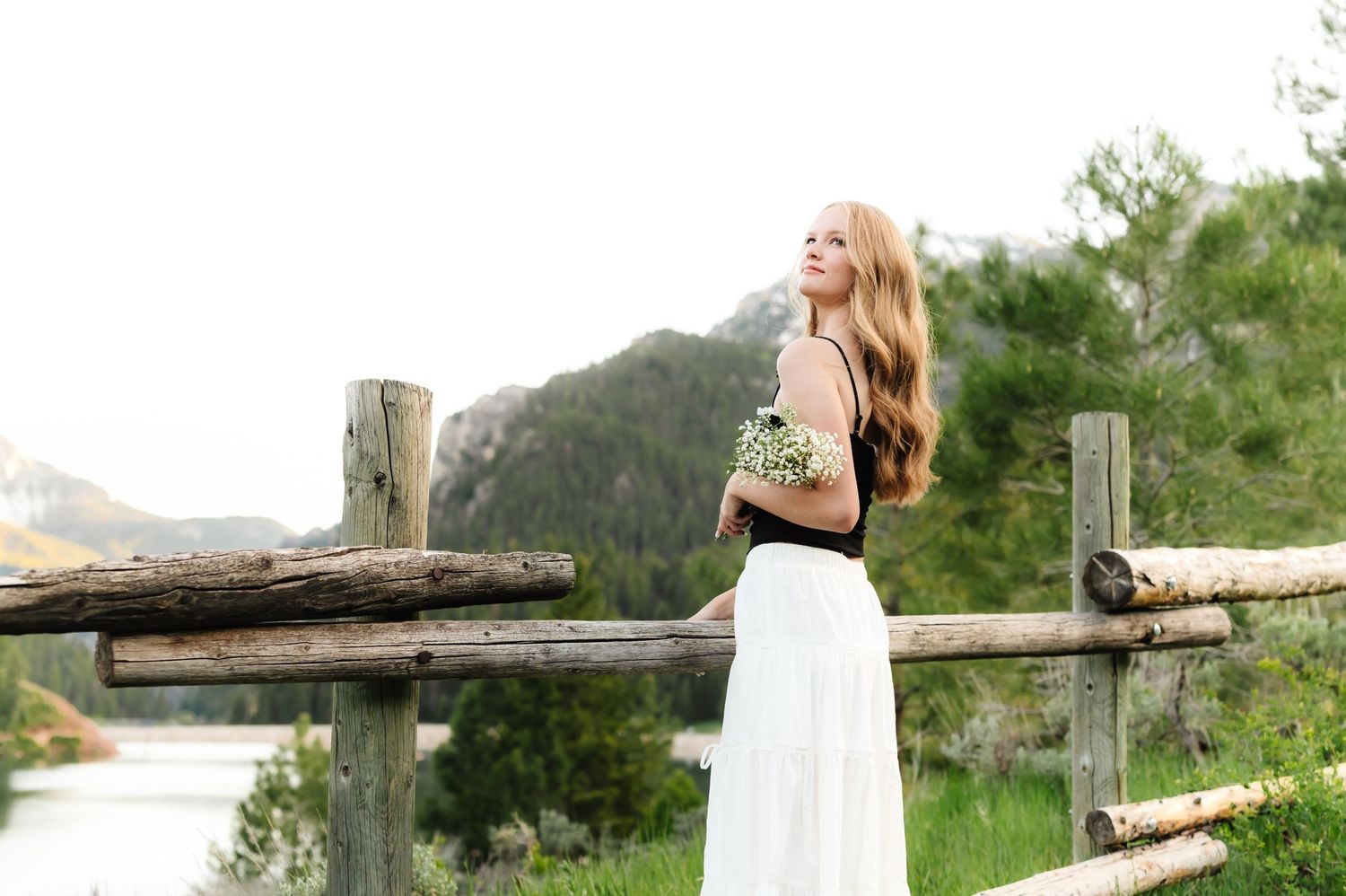 Person in white dress leaning on wooden fence with mountain scenery.