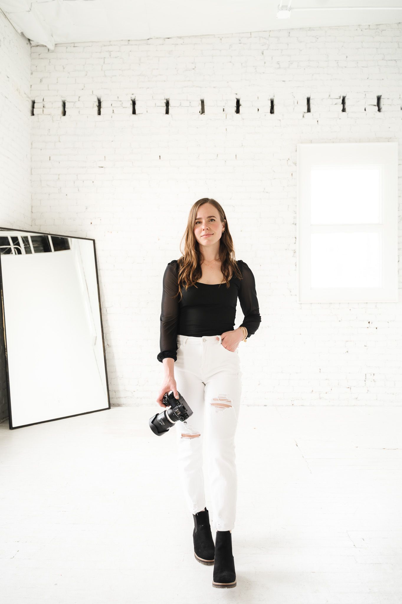 Photographer in black top and white jeans holding camera in minimalist white studio.