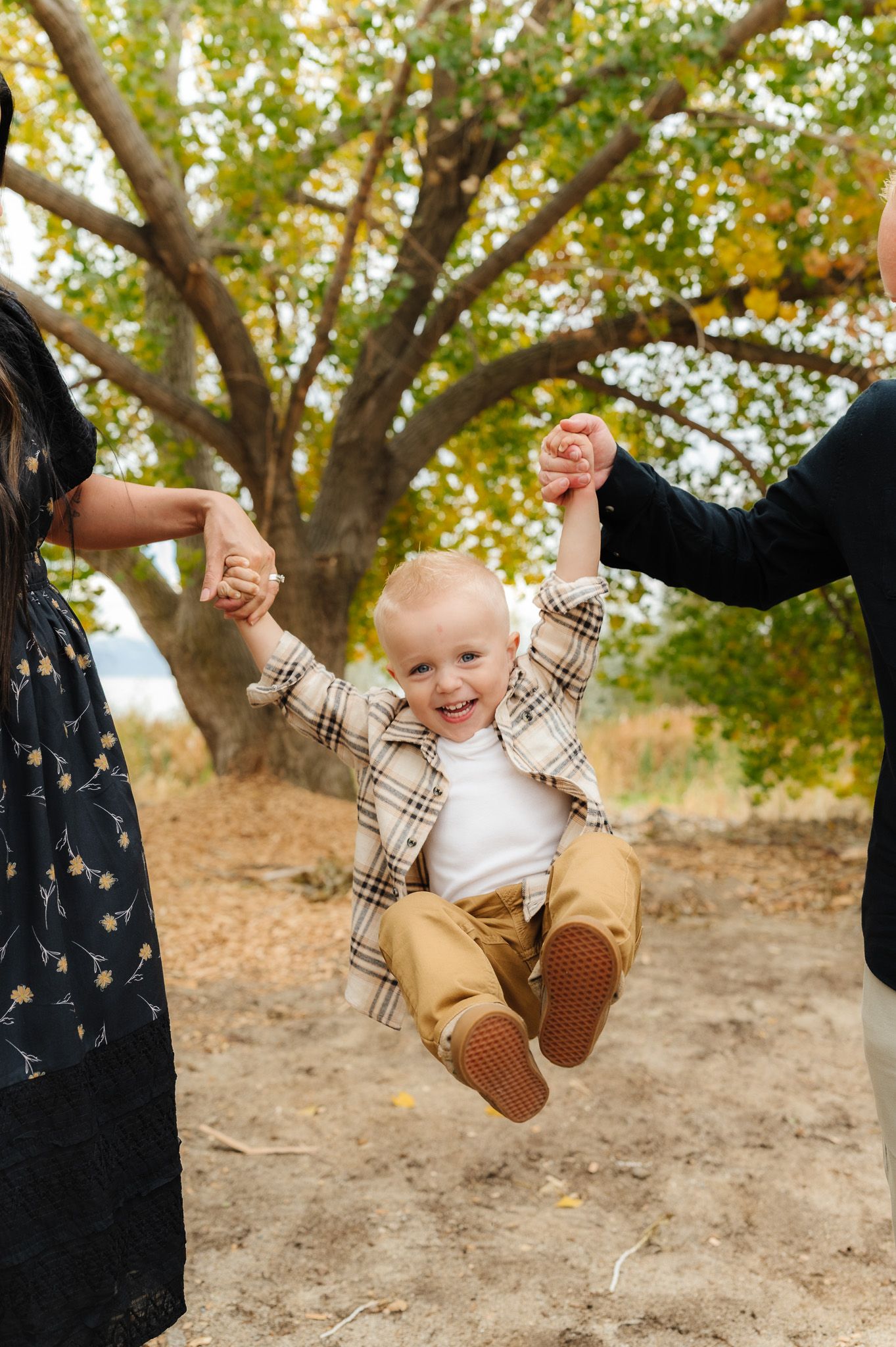 Baby in plaid shirt and khakis being swung joyfully between two people under autumn tree.
