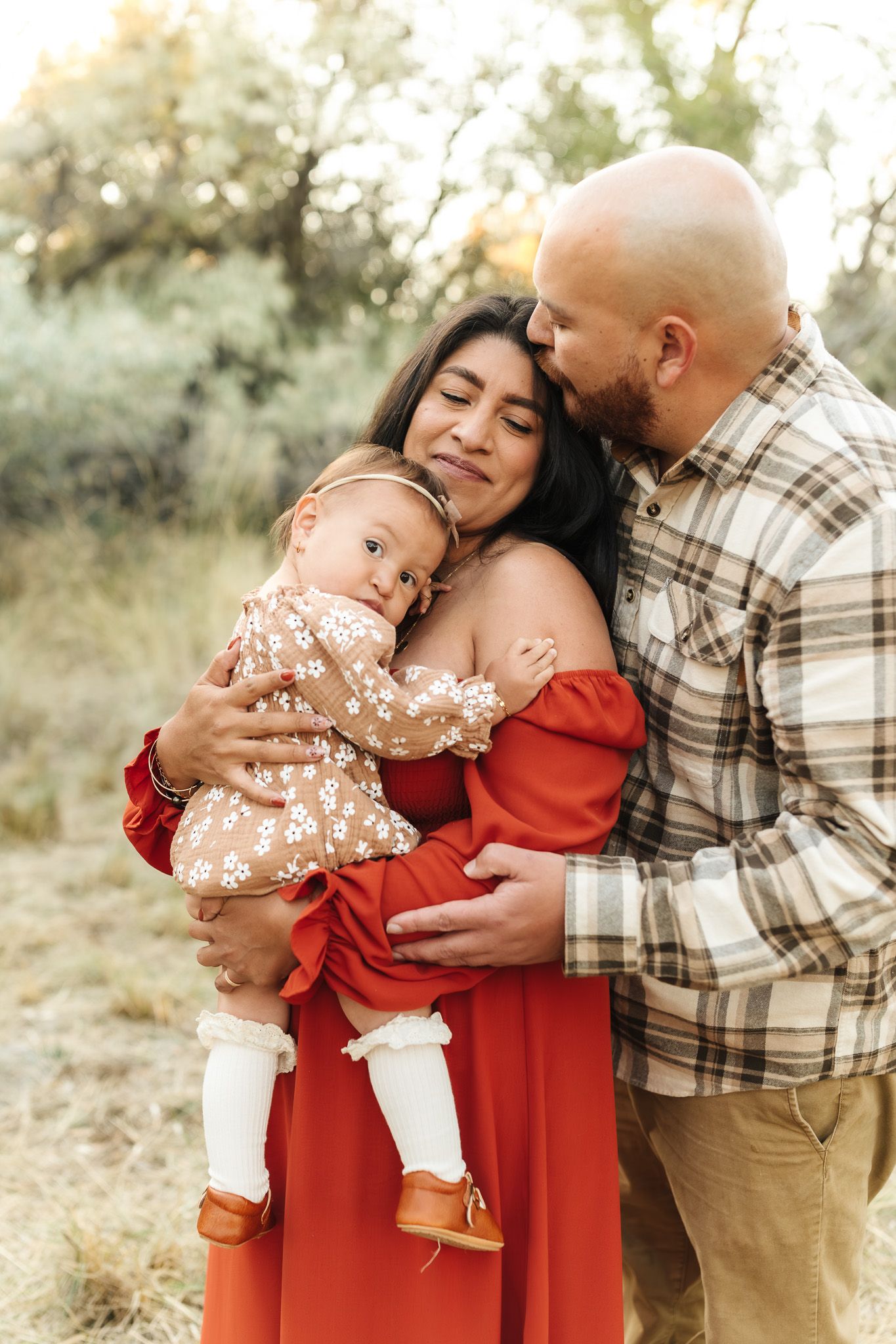 Happy family with bald father kissing brunette mother holding their baby girl outdoors.