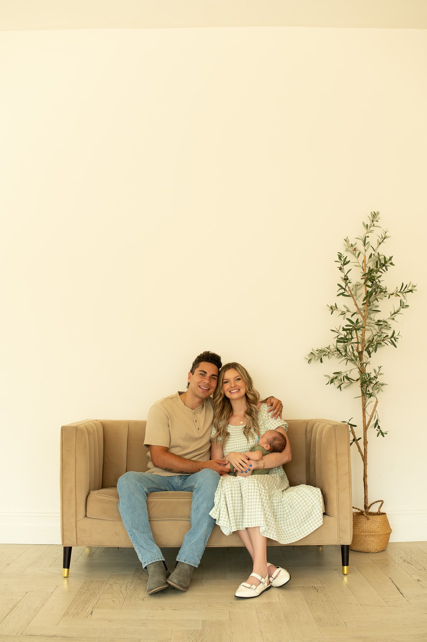 Casual family portrait on beige couch against white wall with potted olive tree.