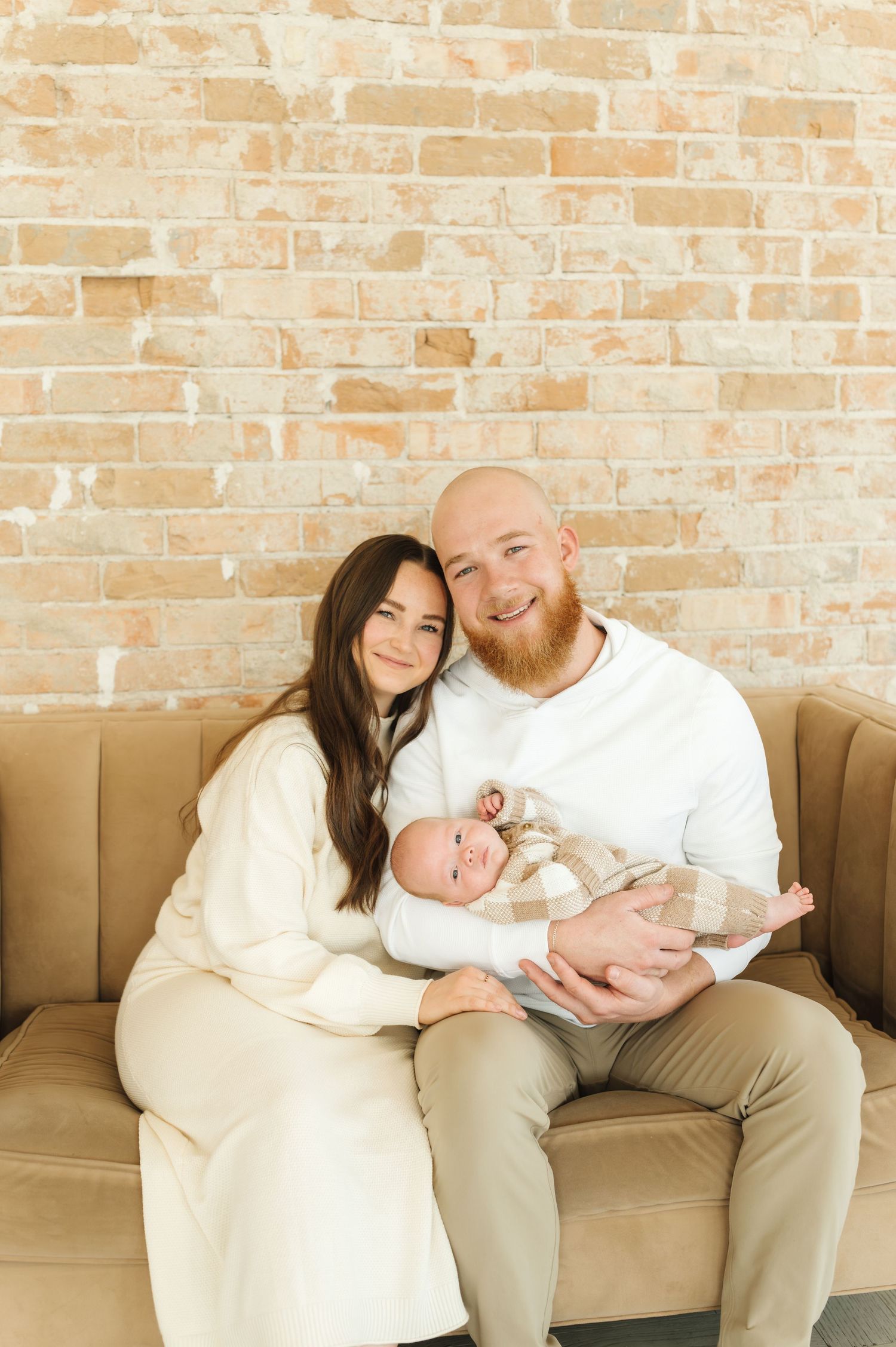 Family portrait on beige couch against exposed brick wall in neutral colored clothing.