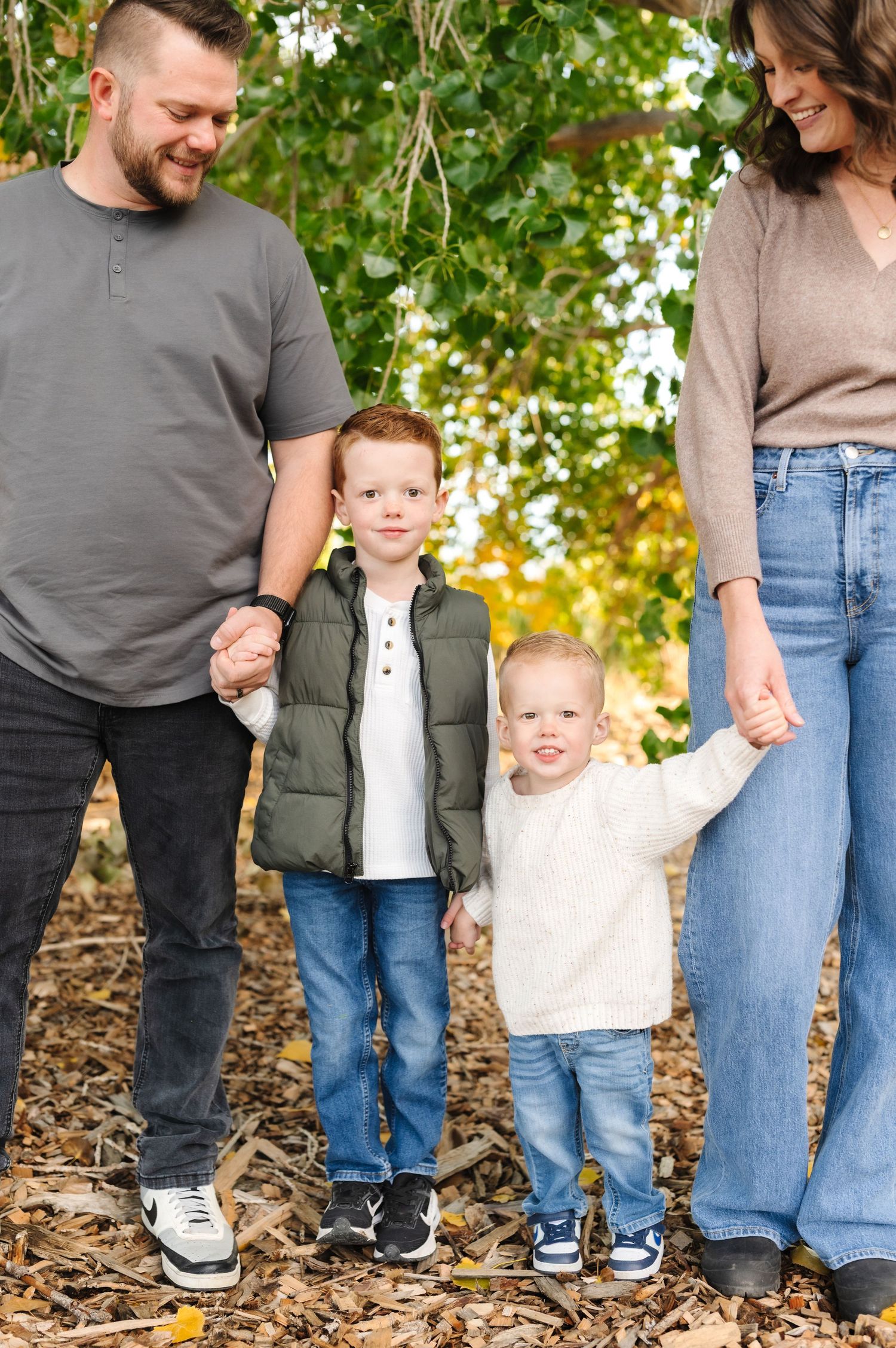 Family of four holding hands outdoors in autumn leaves, two young boys between parents under green trees.