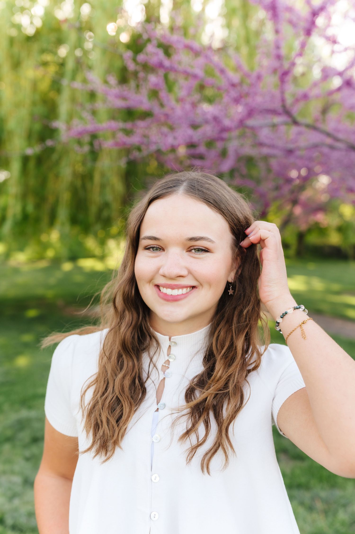 A person in a white blouse poses outdoors with blooming pink trees and greenery in the background.