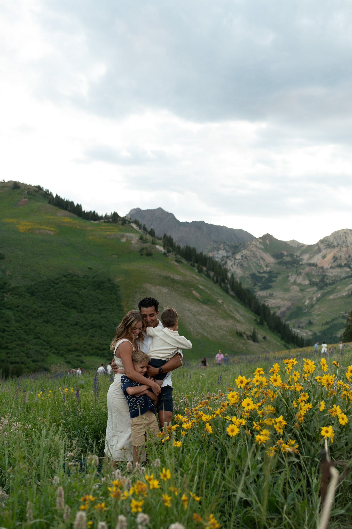 Family with two children posing among yellow wildflowers in a lush mountain meadow under cloudy skies.