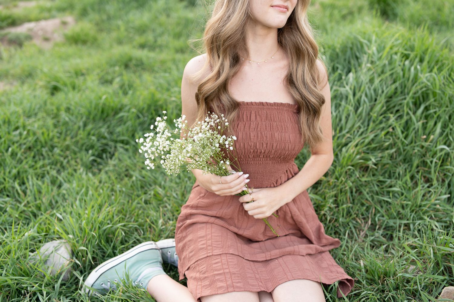 Young woman in rust dress holding white wildflowers, sitting in lush green grass outdoors.