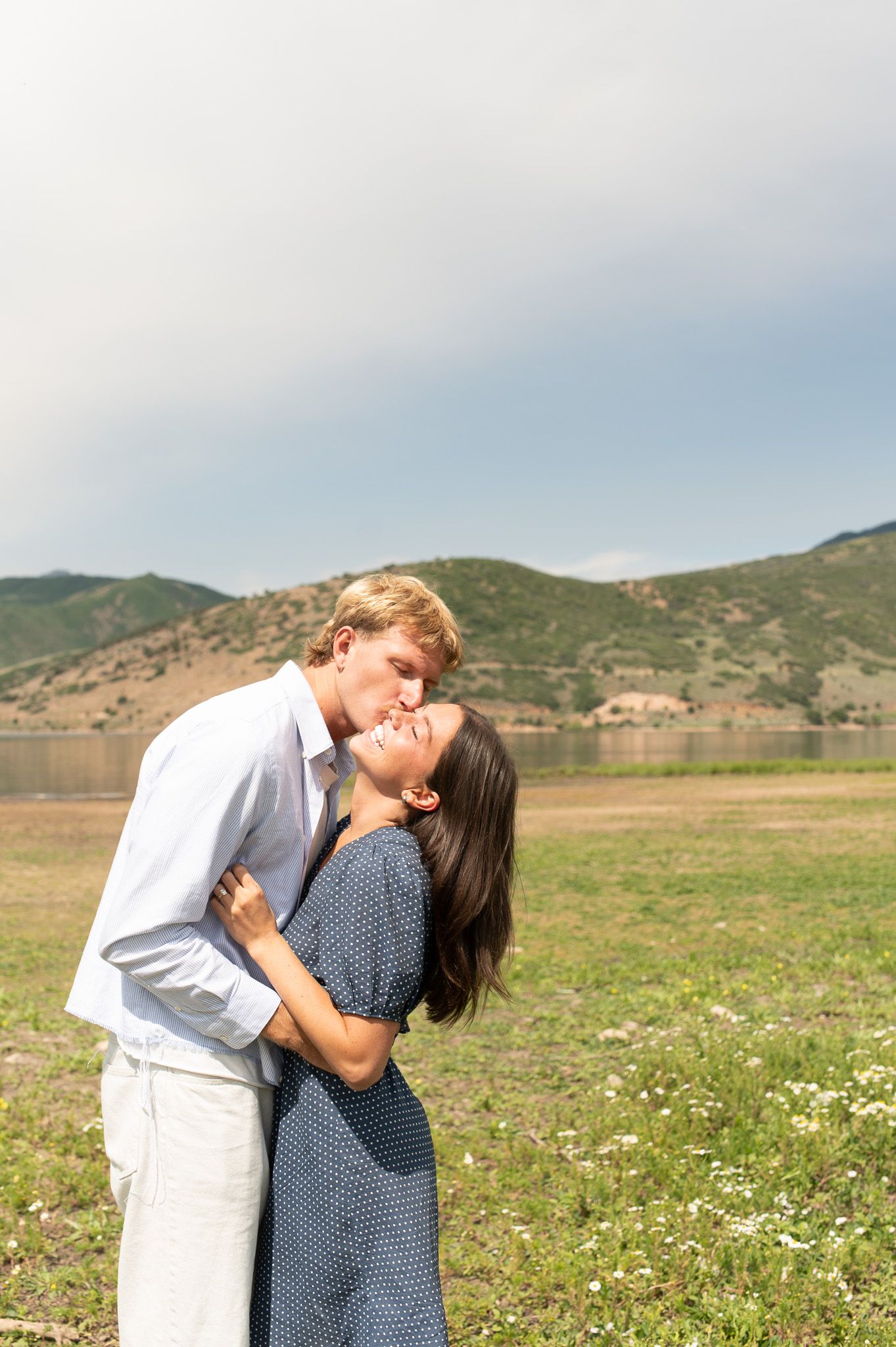 Couple sharing a romantic kiss outdoors near a lake with green hills and cloudy sky in background.