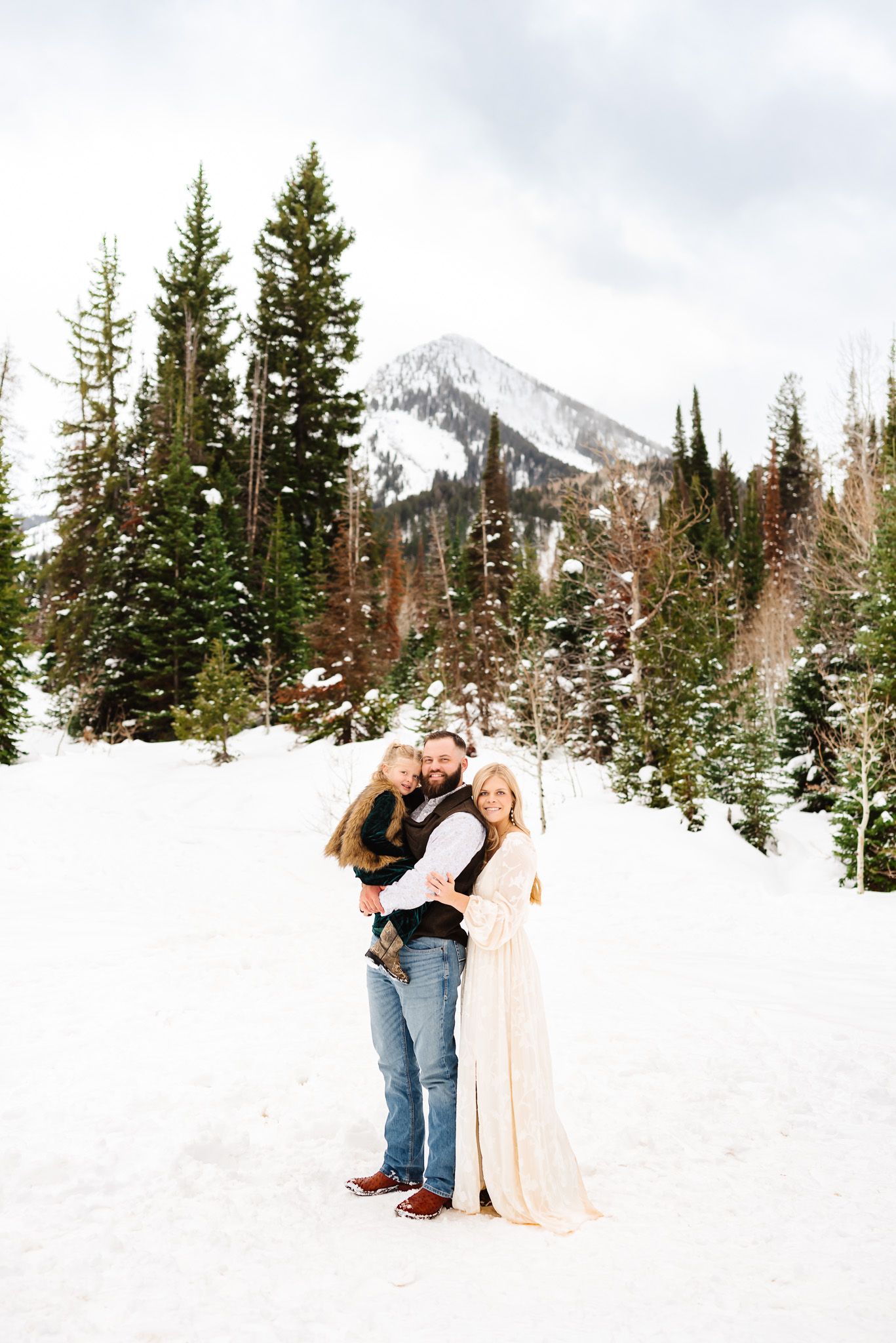 Romantic winter photoshoot with snow-capped mountains and pine trees in the background.