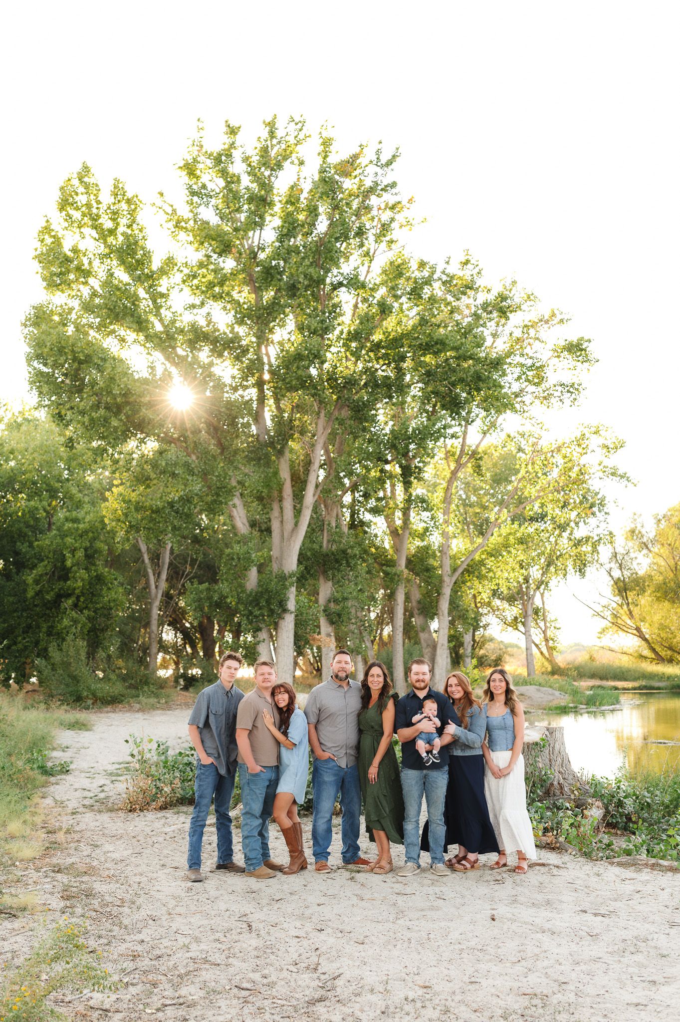 Large family group portrait outdoors near a river with lush green trees and golden sunset light.