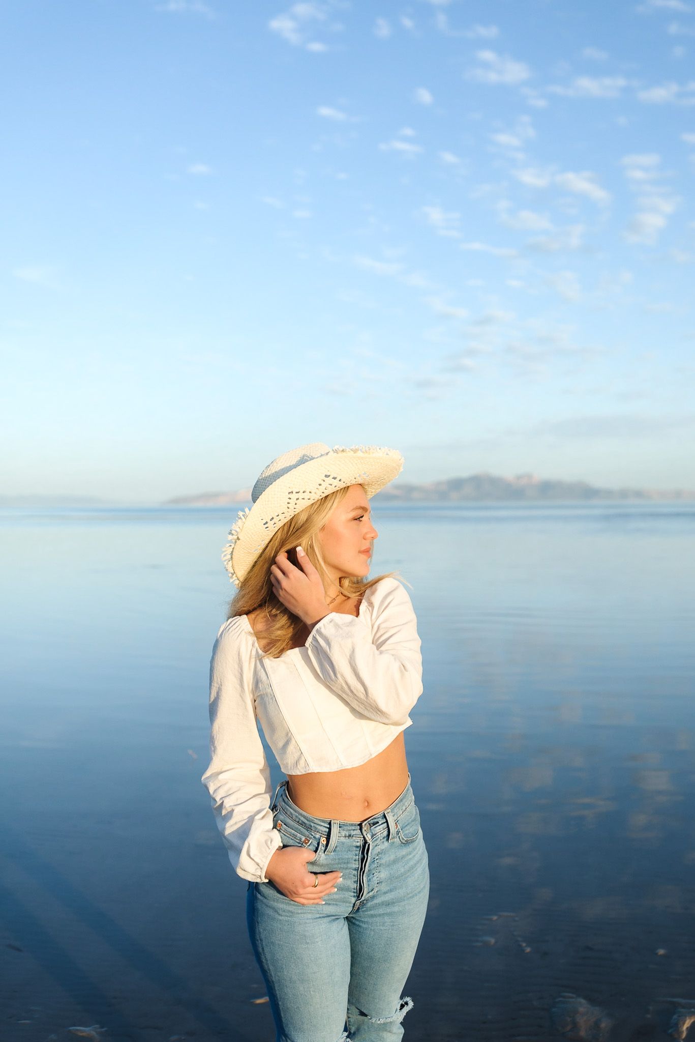 Blonde woman wearing straw hat and white crop top stands near serene lake with mountain backdrop at sunset.