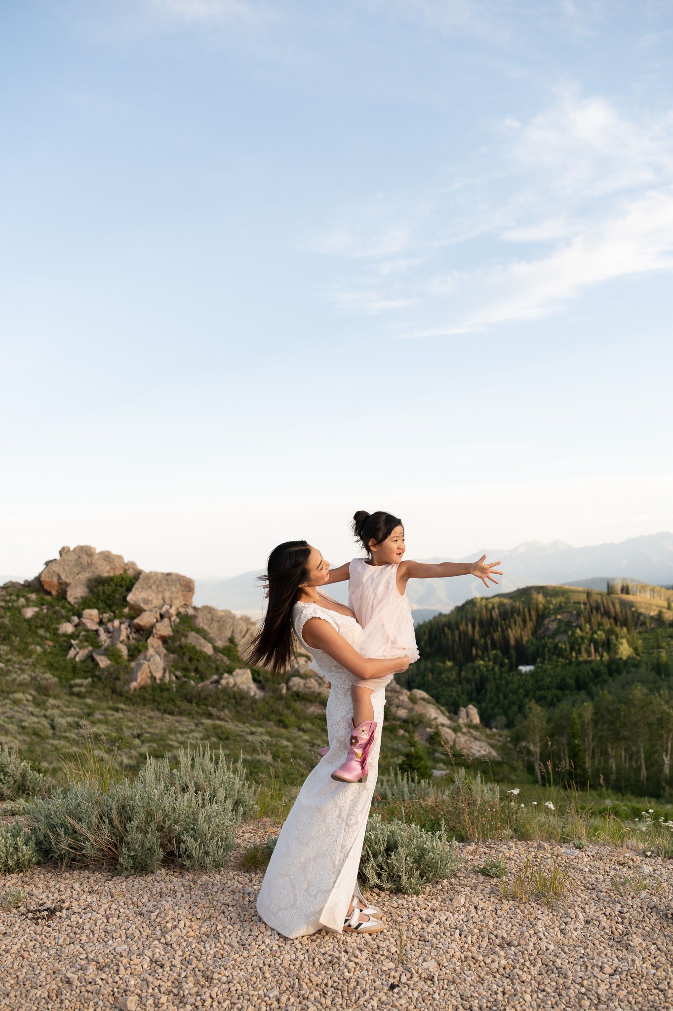A mother lifts her young daughter outdoors, both in white dresses, with mountains and blue sky behind them.