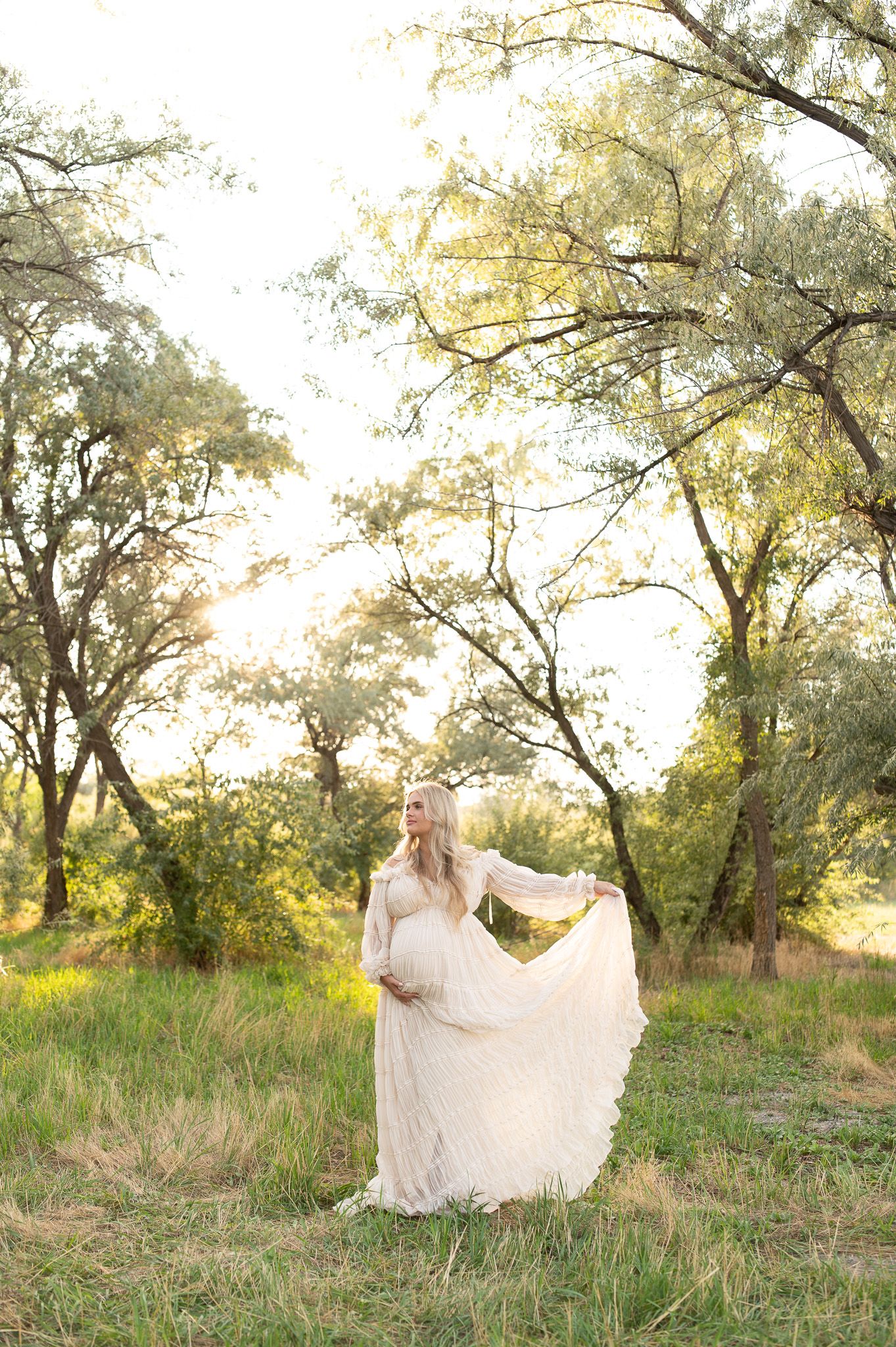 Pregnant woman in flowing white lace gown standing in sunlit meadow surrounded by trees during golden hour maternity photoshoot.
