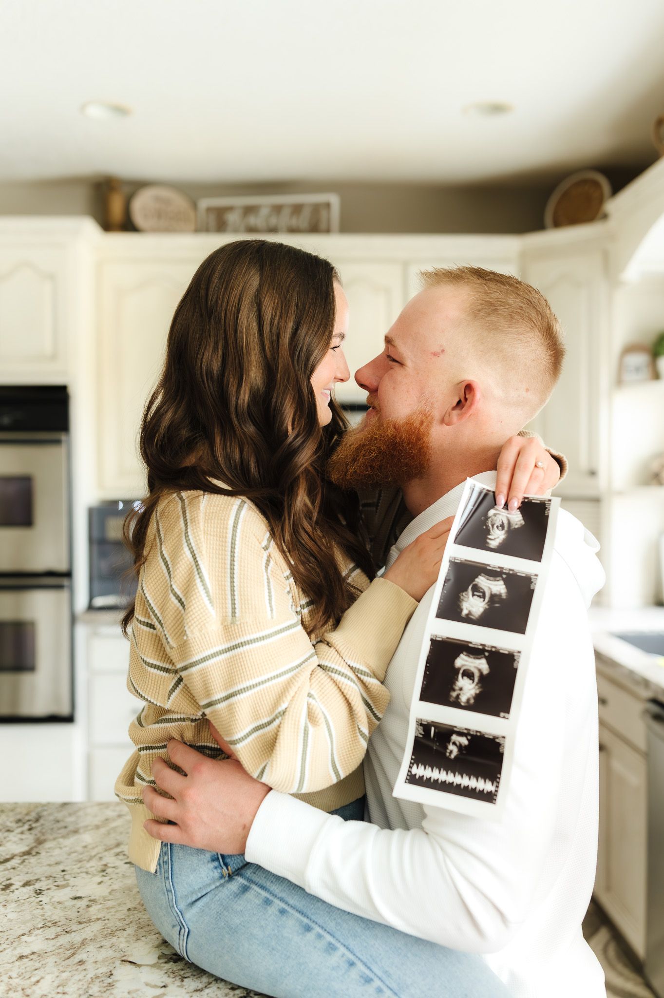 A couple shares a romantic moment in a kitchen, holding ultrasound photos, announcing their pregnancy.