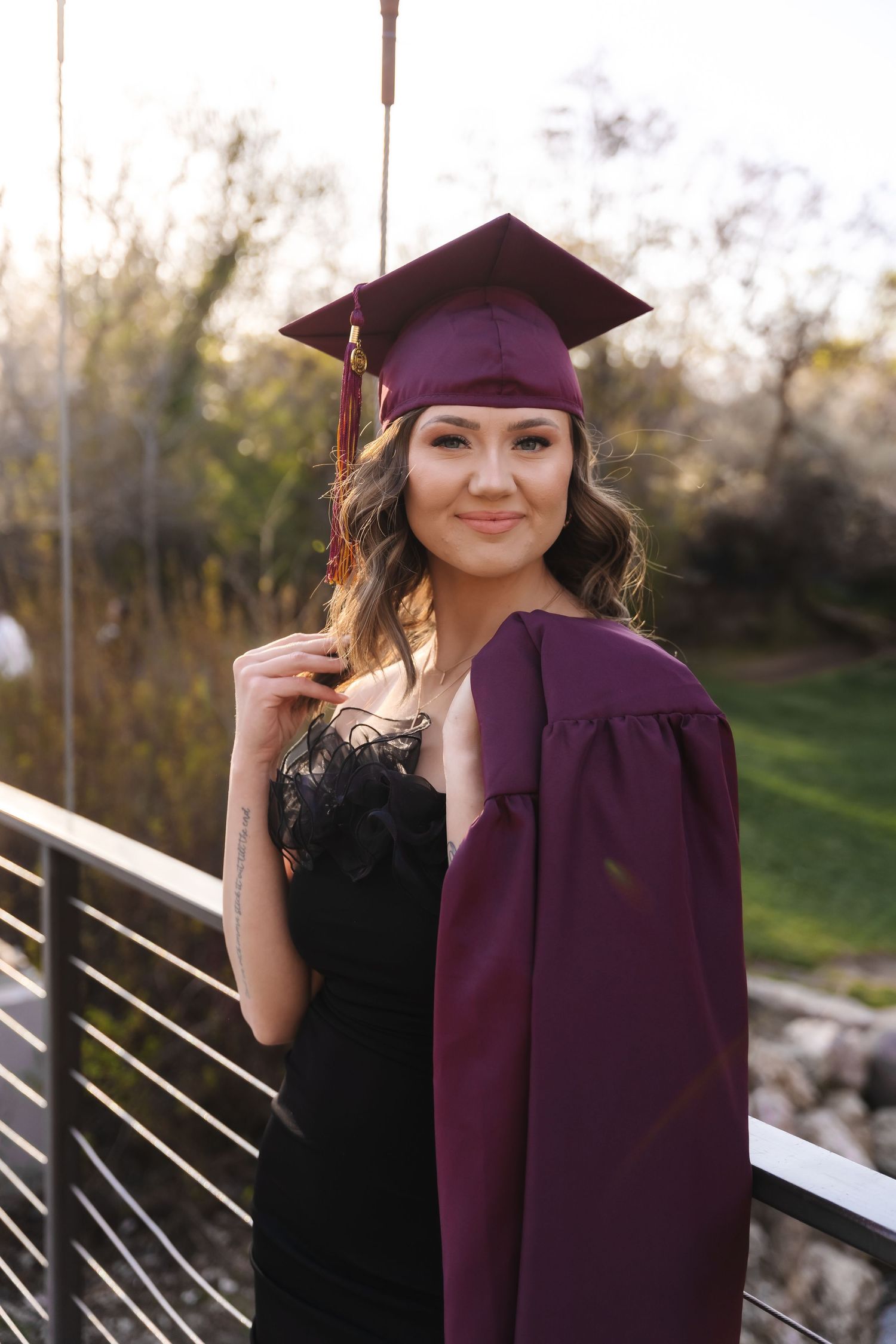 Graduate in black dress poses with maroon cap and gown against outdoor backdrop.