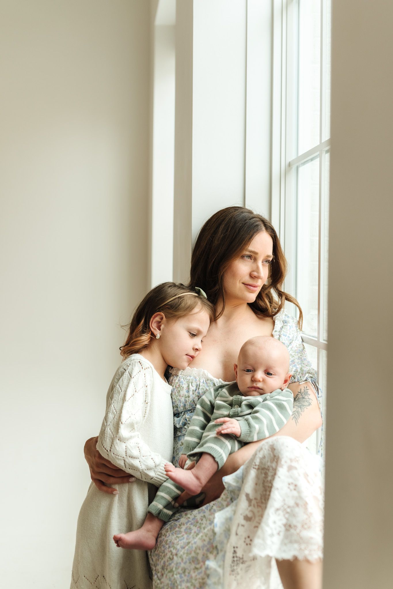 A mother holds her baby and embraces her young daughter by a bright window in soft natural light.