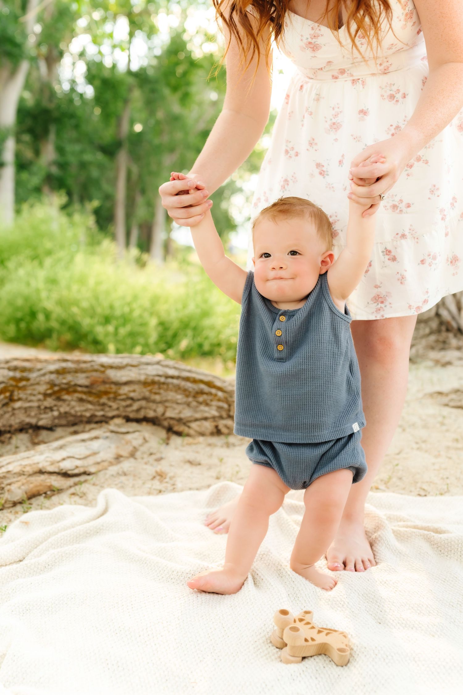 Baby in blue romper sitting on white blanket in outdoor setting with natural lighting.