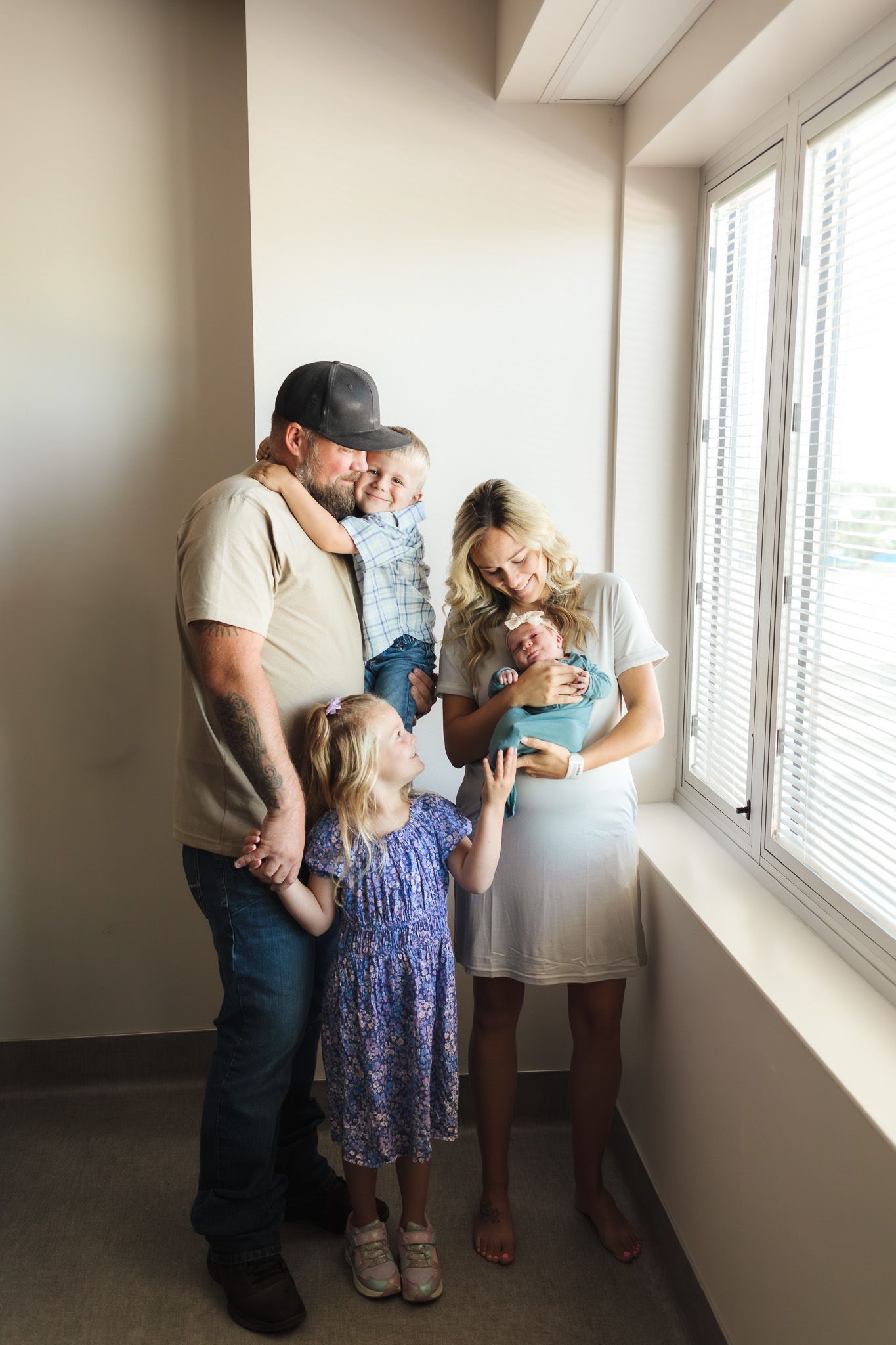 Family of four with newborn baby gathered by hospital window, parents holding children in tender moment.