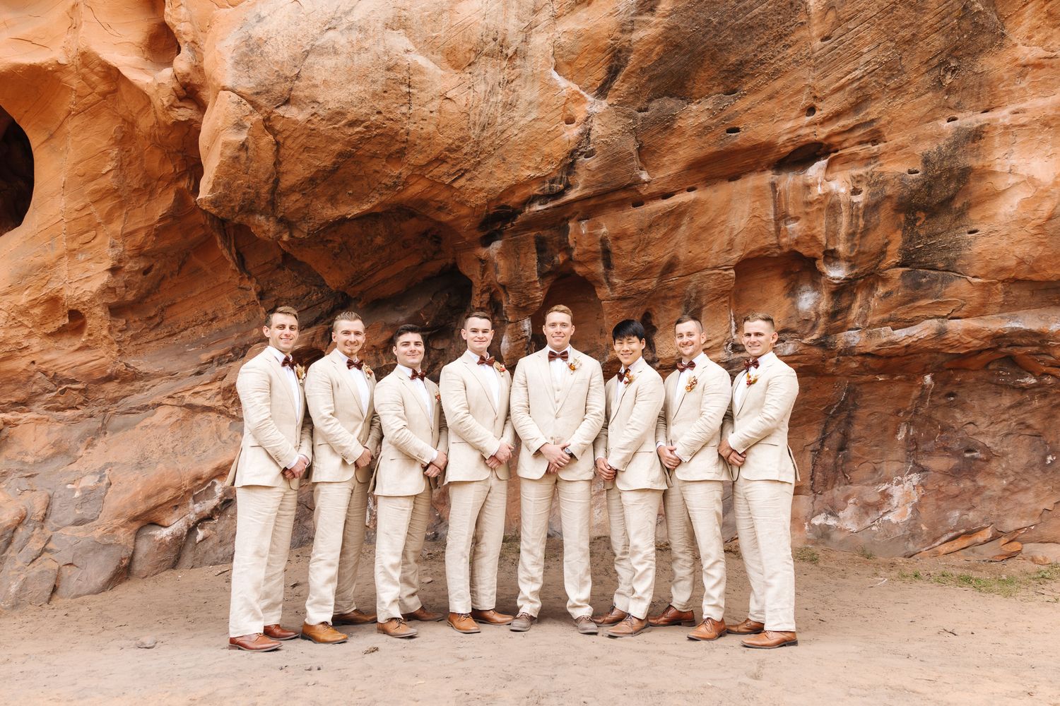 Groomsmen in tan suits with burgundy boutonnieres posing in front of red rock canyon walls at an outdoor wedding.