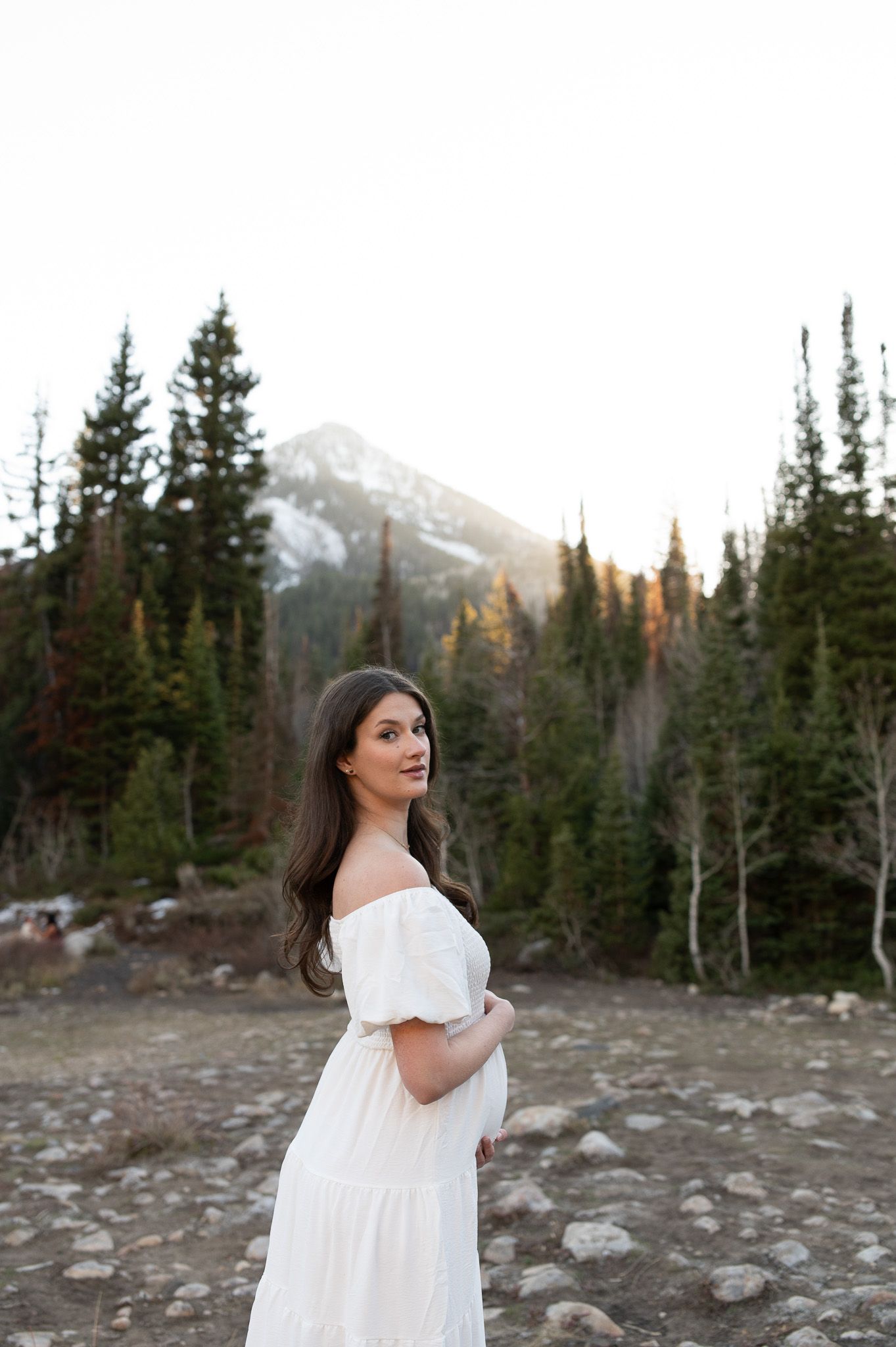 Woman in white off-shoulder dress poses in rocky mountain landscape with pine trees and snow-capped peak at sunset.