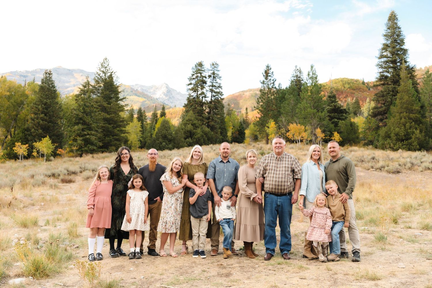 Large family posing outdoors in a mountain meadow surrounded by pine trees and fall foliage.