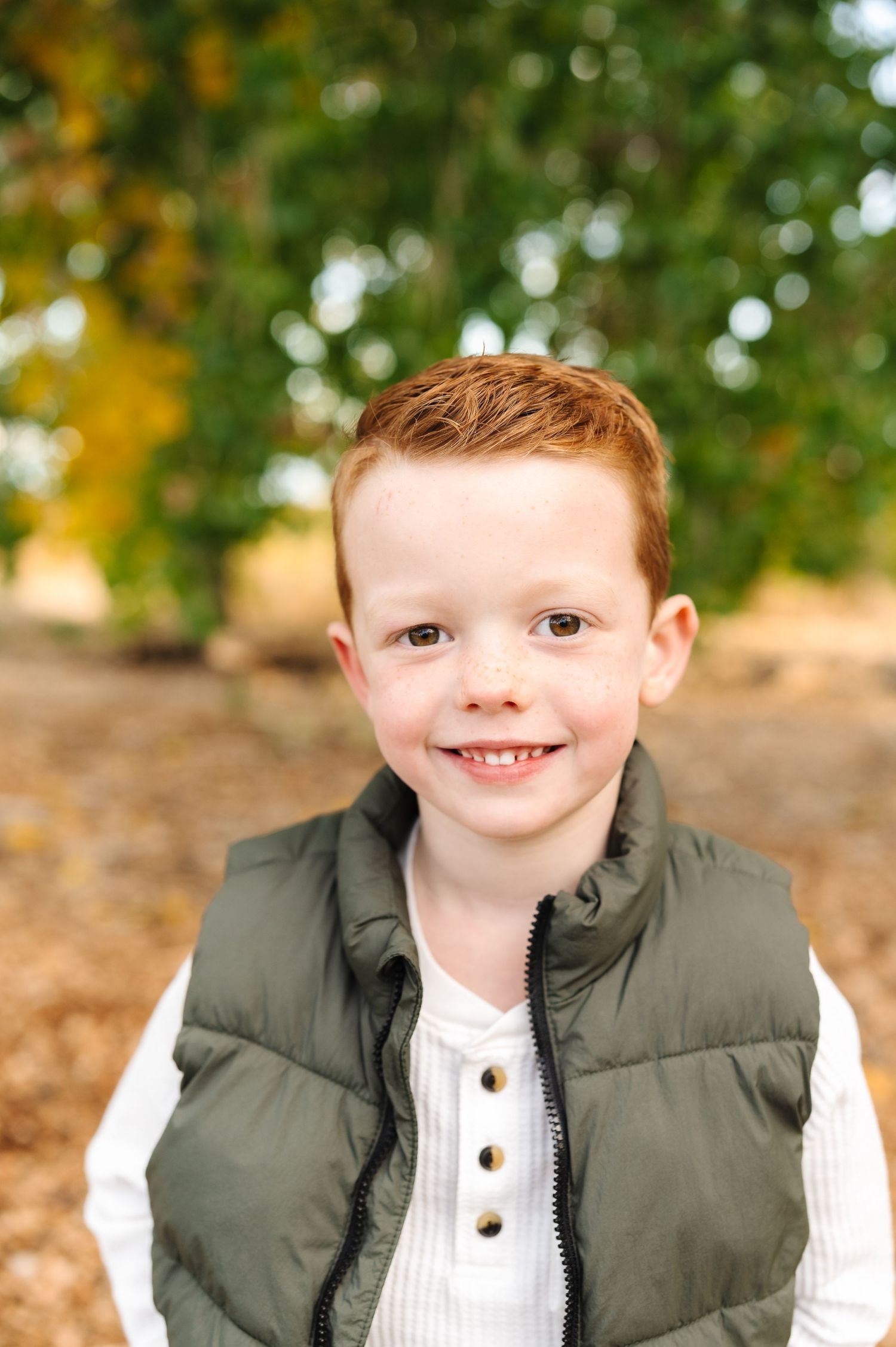 Smiling red-haired boy wearing a green vest and white shirt outdoors in a fall setting.