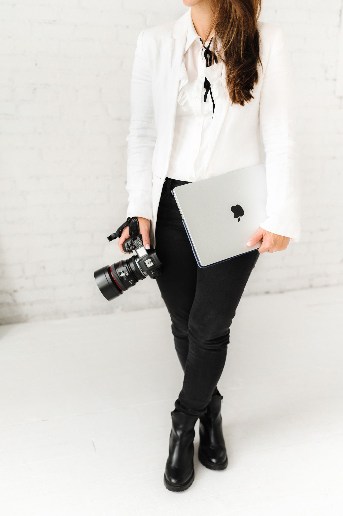 Professional photographer wearing white blazer and black pants holds camera in studio setting.