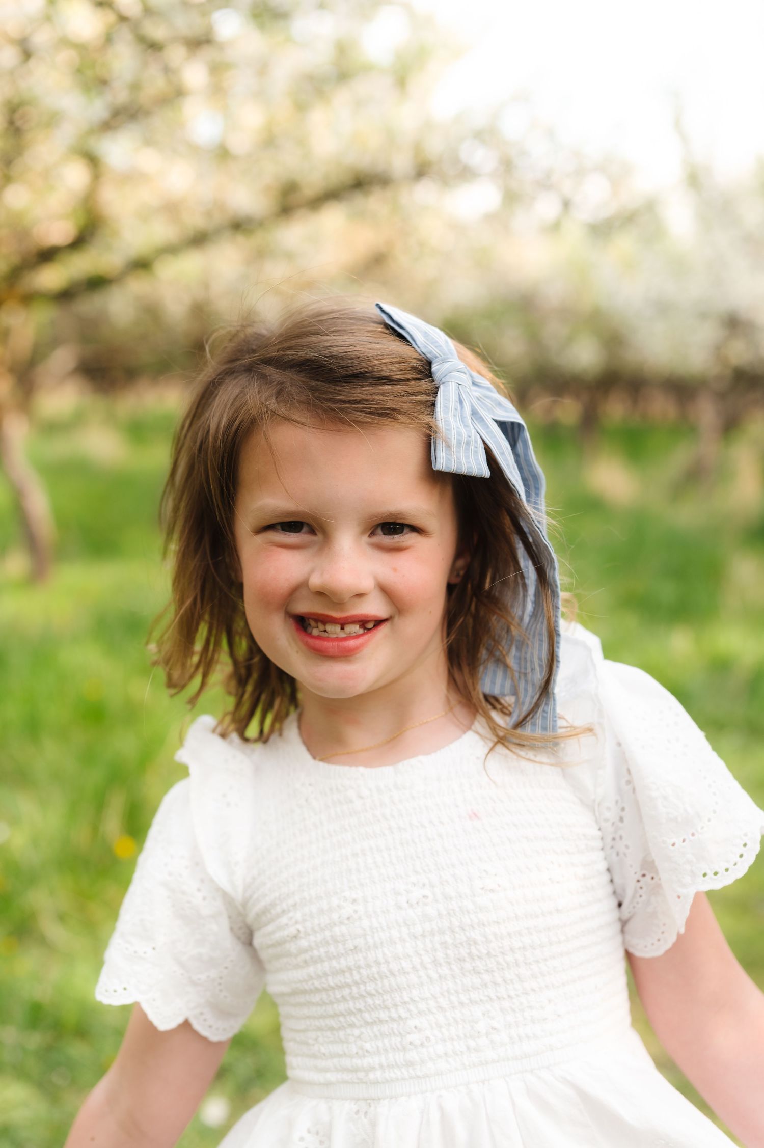 A smiling child wearing a white lace dress and blue hair bow stands in a spring orchard setting.