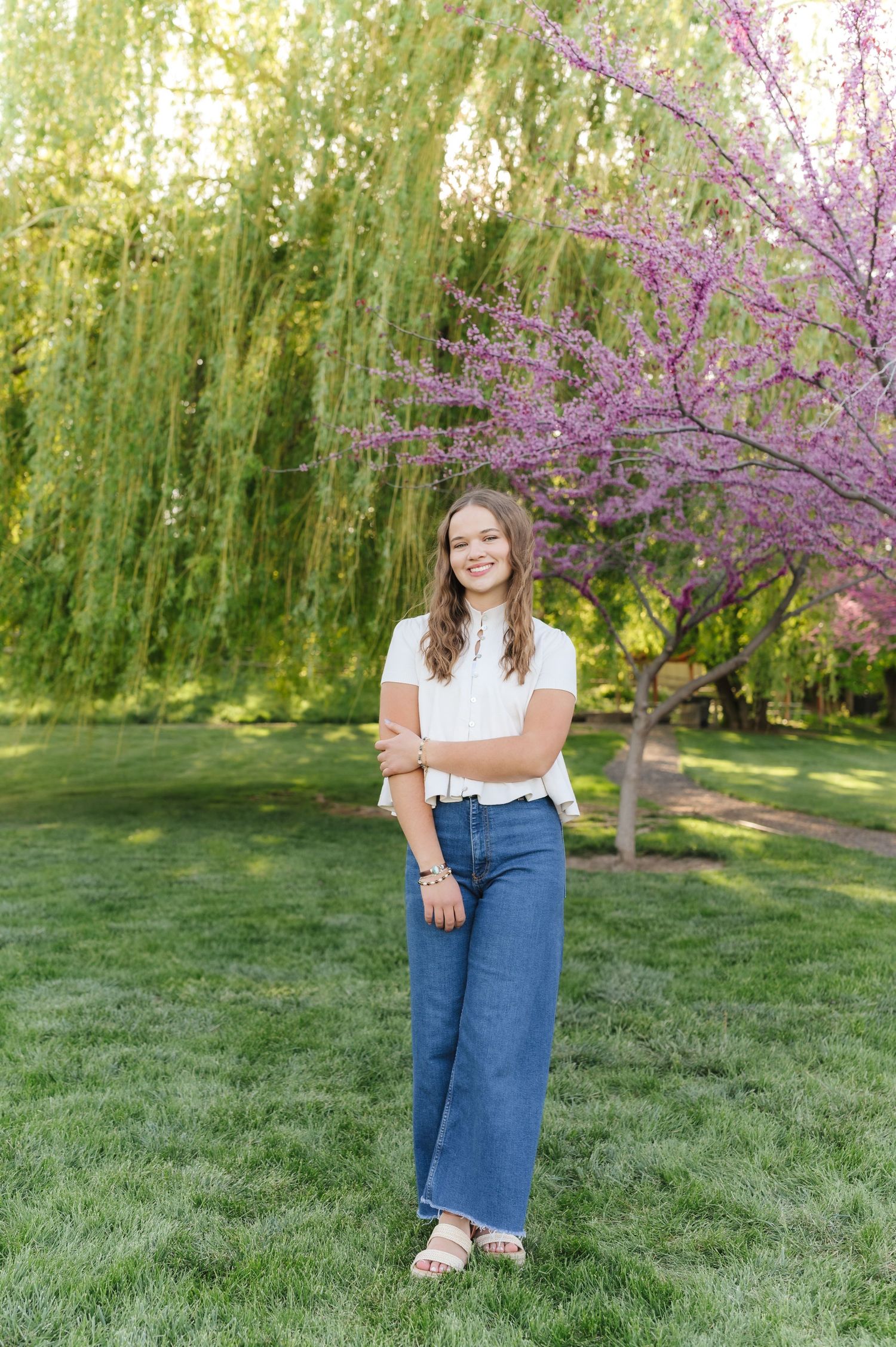 Person in white top and denim skirt poses on grass with blooming cherry trees and weeping willows behind.