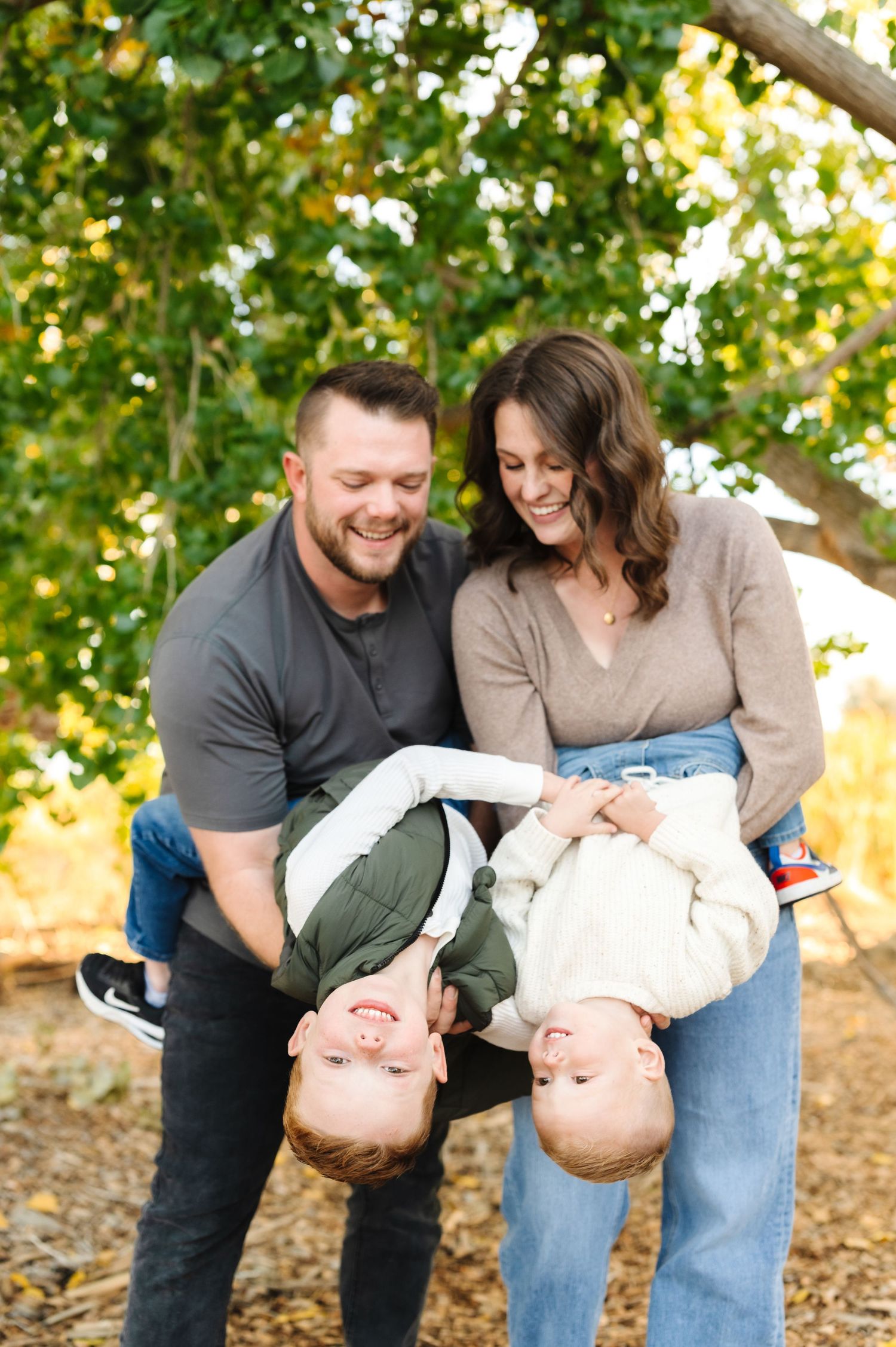 A happy family of four with two young children being held upside down by their smiling parents outdoors under green trees.