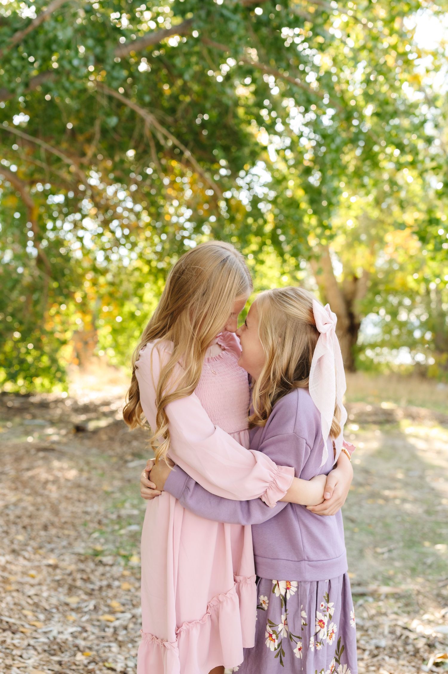Two young girls in pink and purple dresses hugging and touching foreheads outdoors among green trees.