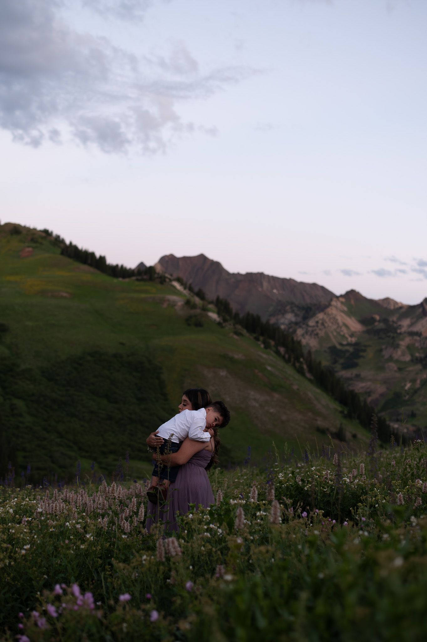 Couple embracing in a wildflower meadow with green mountain slopes and a soft twilight sky in the background.