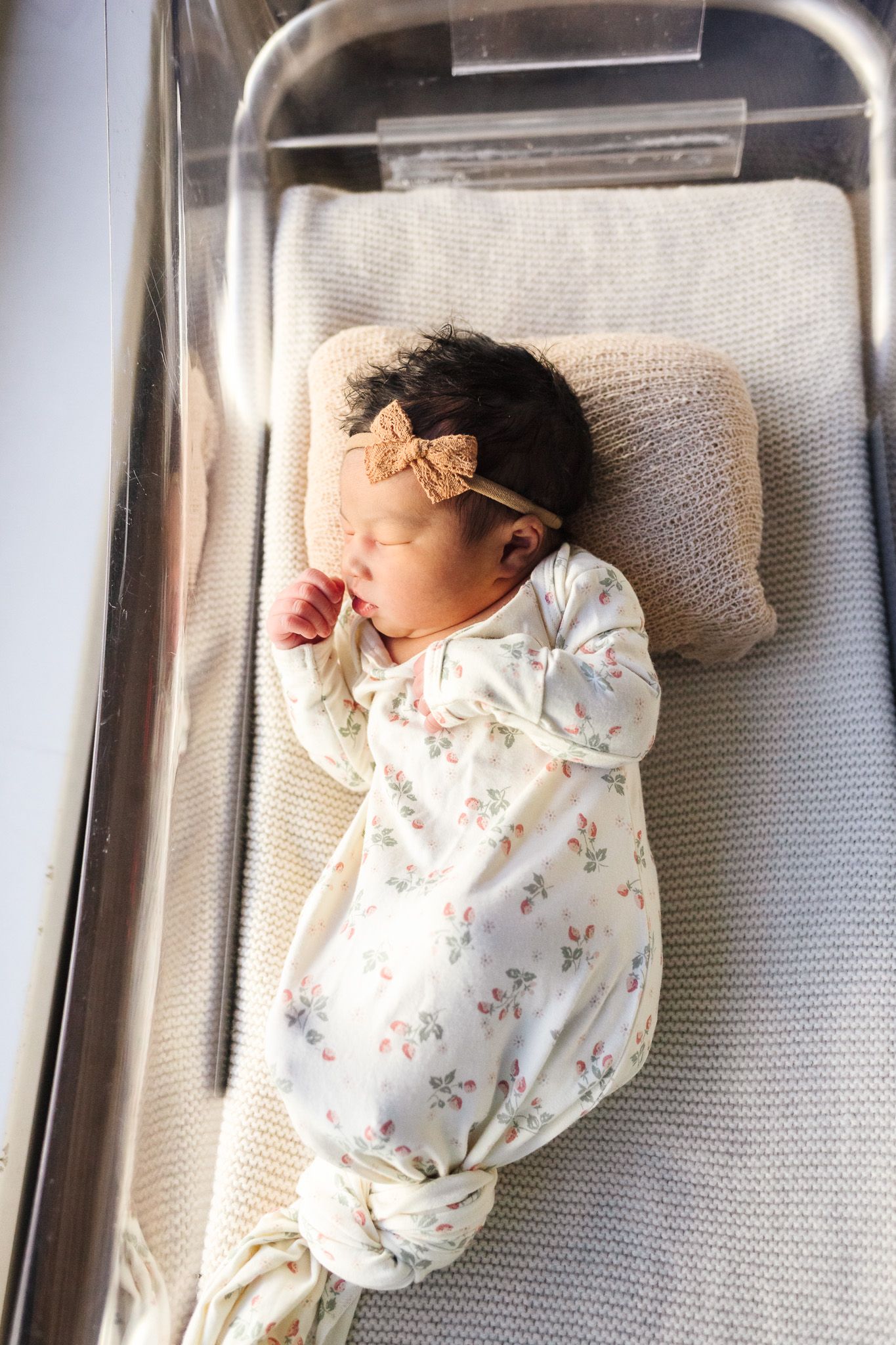 Newborn baby girl sleeping in hospital bassinet wearing floral onesie and bow headband.