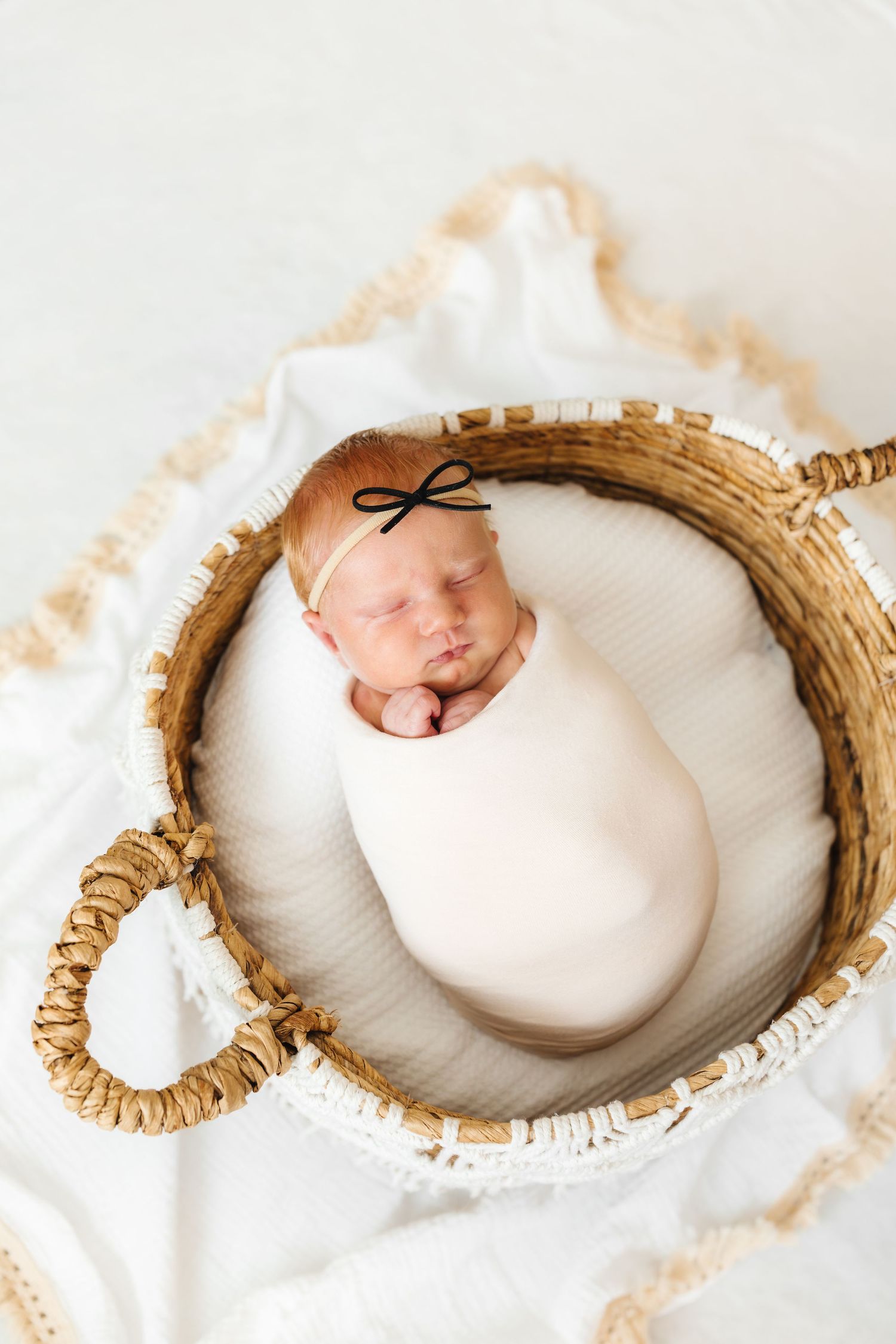 Newborn baby swaddled in white wrap sleeping peacefully in a wicker basket with a small black bow headband.