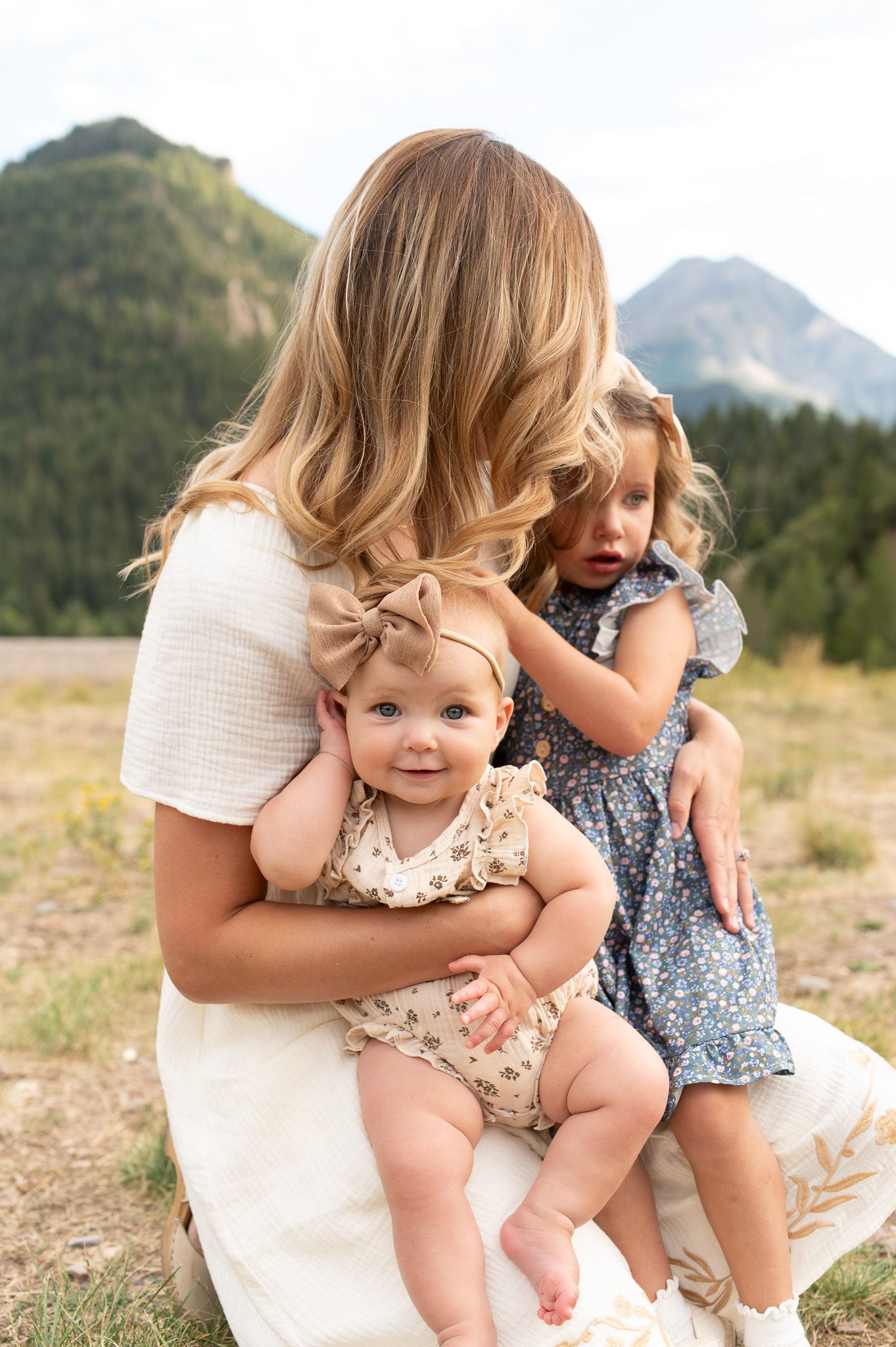 Mother holding two young daughters in floral outfits with mountain landscape in background.