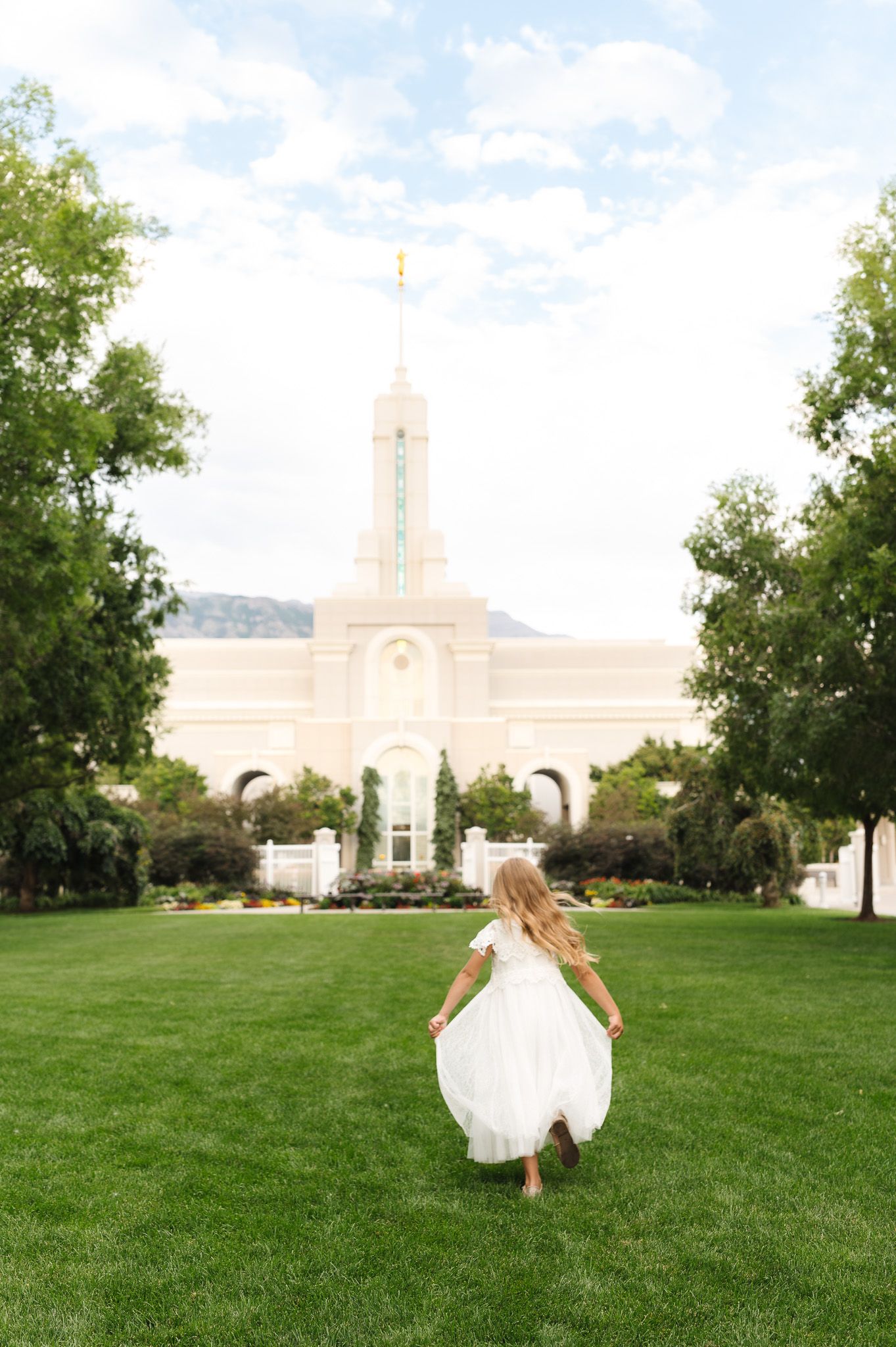 Young girl in white dress running toward an LDS temple on a sunny day with green lawn and trees.
