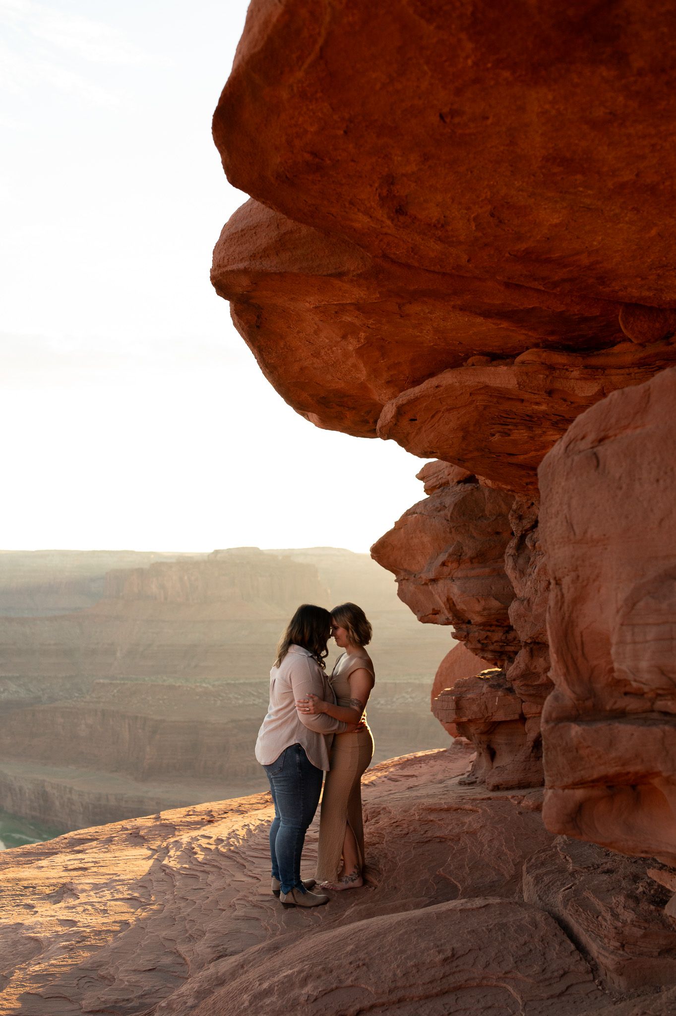 Couple embracing under a red rock overhang at sunset with a vast canyon landscape in the background.