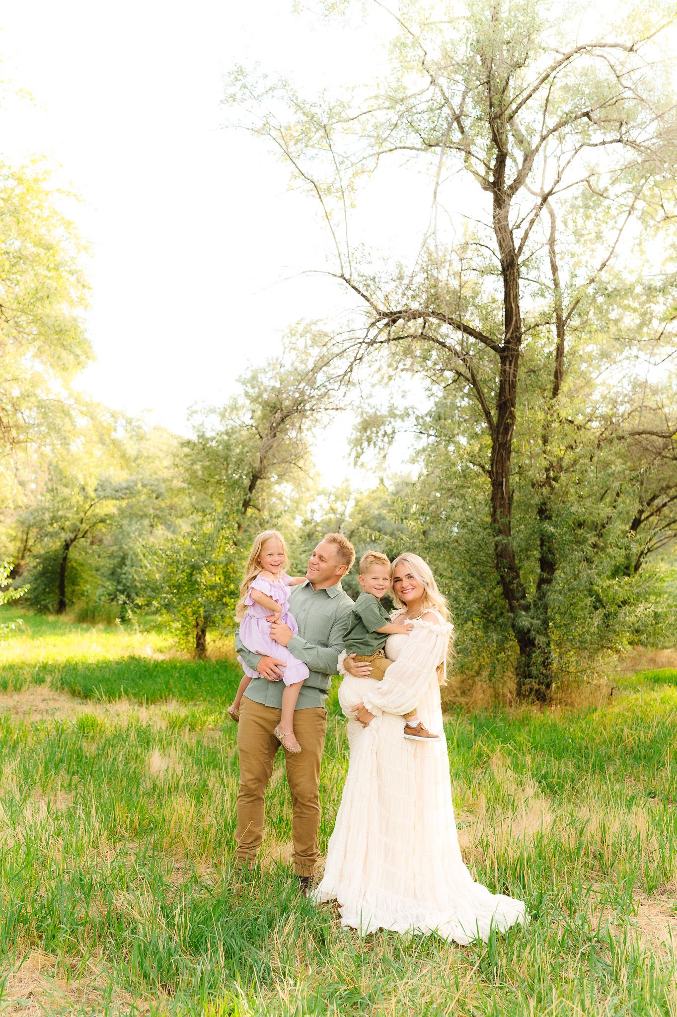 Family portrait session in a sunlit meadow with tall grass and trees.