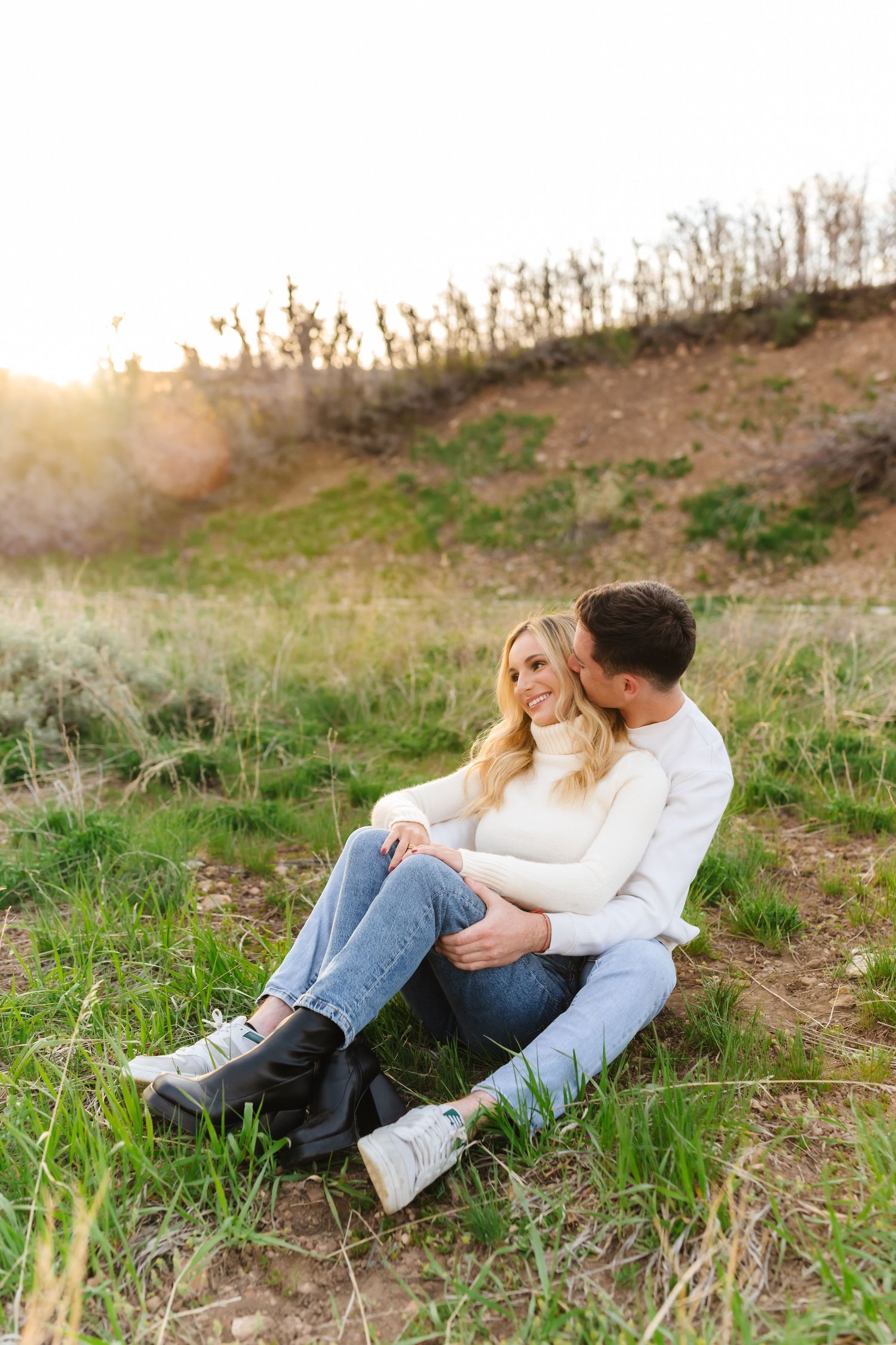 Couple in casual attire sharing intimate moment while sitting in grassy hillside.