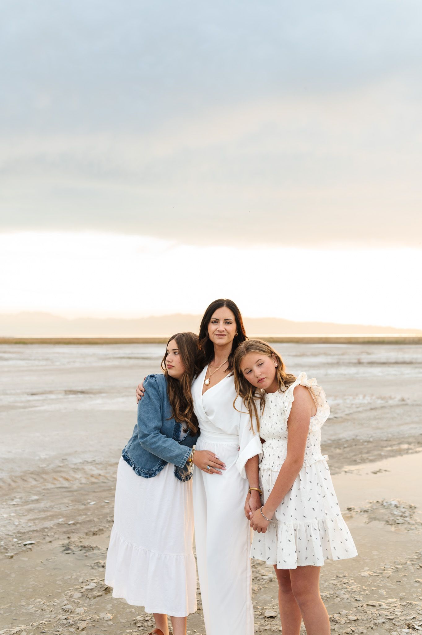 Mother in white dress with two daughters leaning on her at a serene lakeside during golden hour.