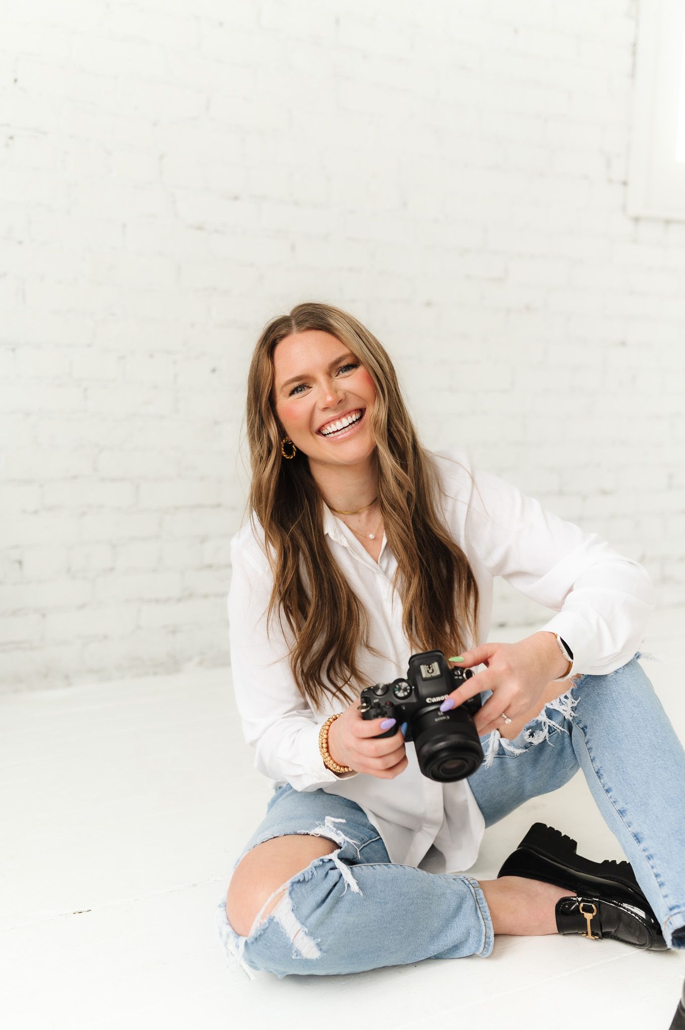 Photographer in white shirt and ripped jeans sits casually with camera against white background.