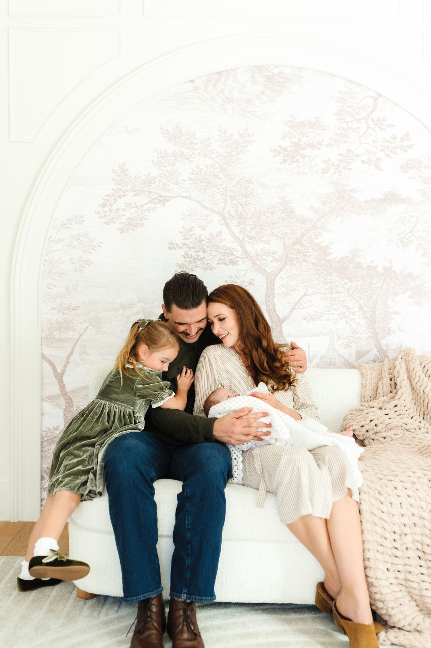 A family of four sits together on a white chair, parents lovingly gazing at their newborn baby as a toddler leans in.