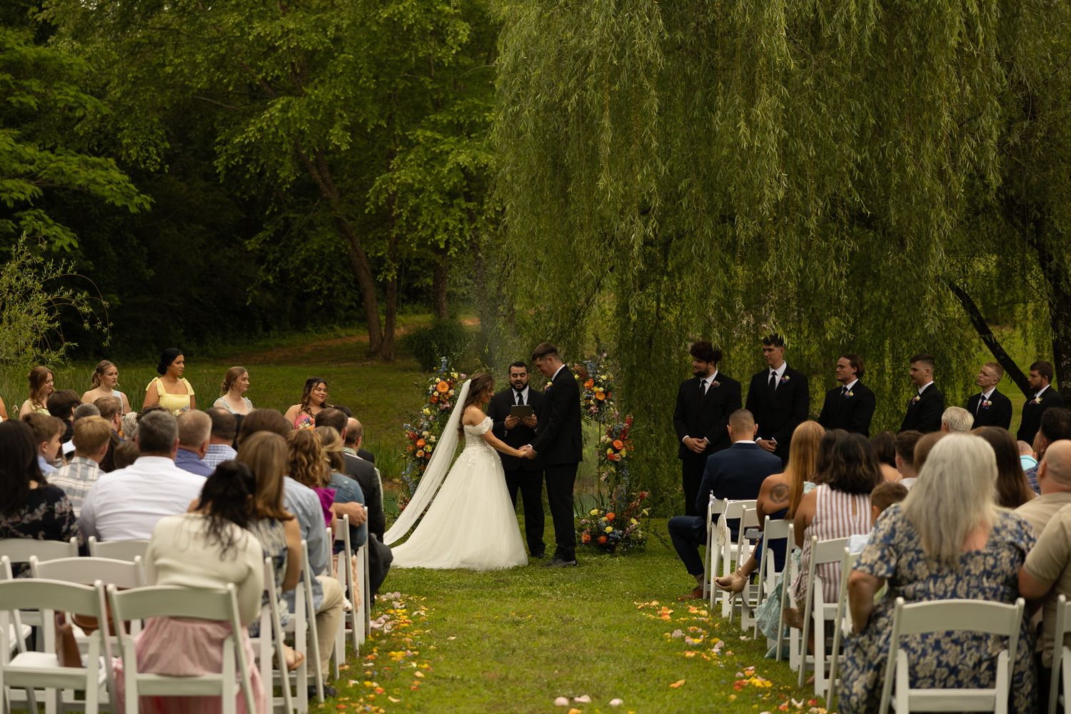 Outdoor wedding ceremony under a weeping willow tree with bride, groom, officiant, and guests seated in white chairs.